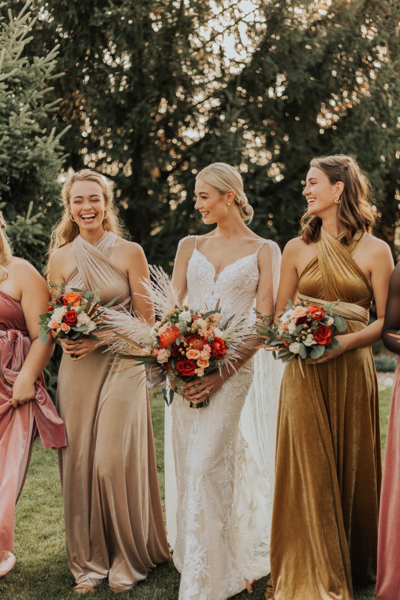 A bride laughs with her bridesmaids at golden hour, all holding bouquets in dresses of terracotta, honey and rose