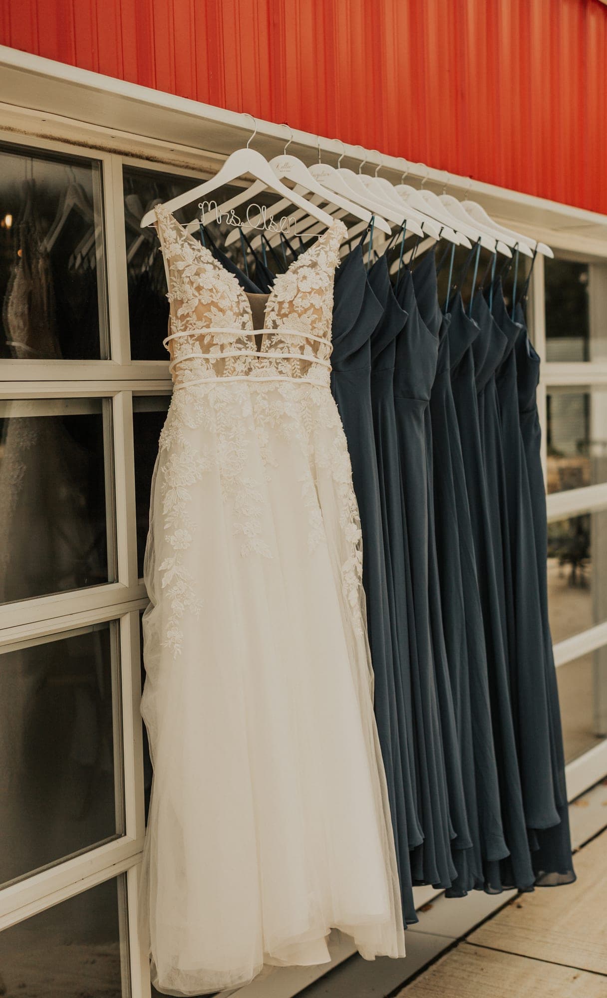 A lace wedding gown hangs beside a row of navy bridesmaid dresses against a red barn wall