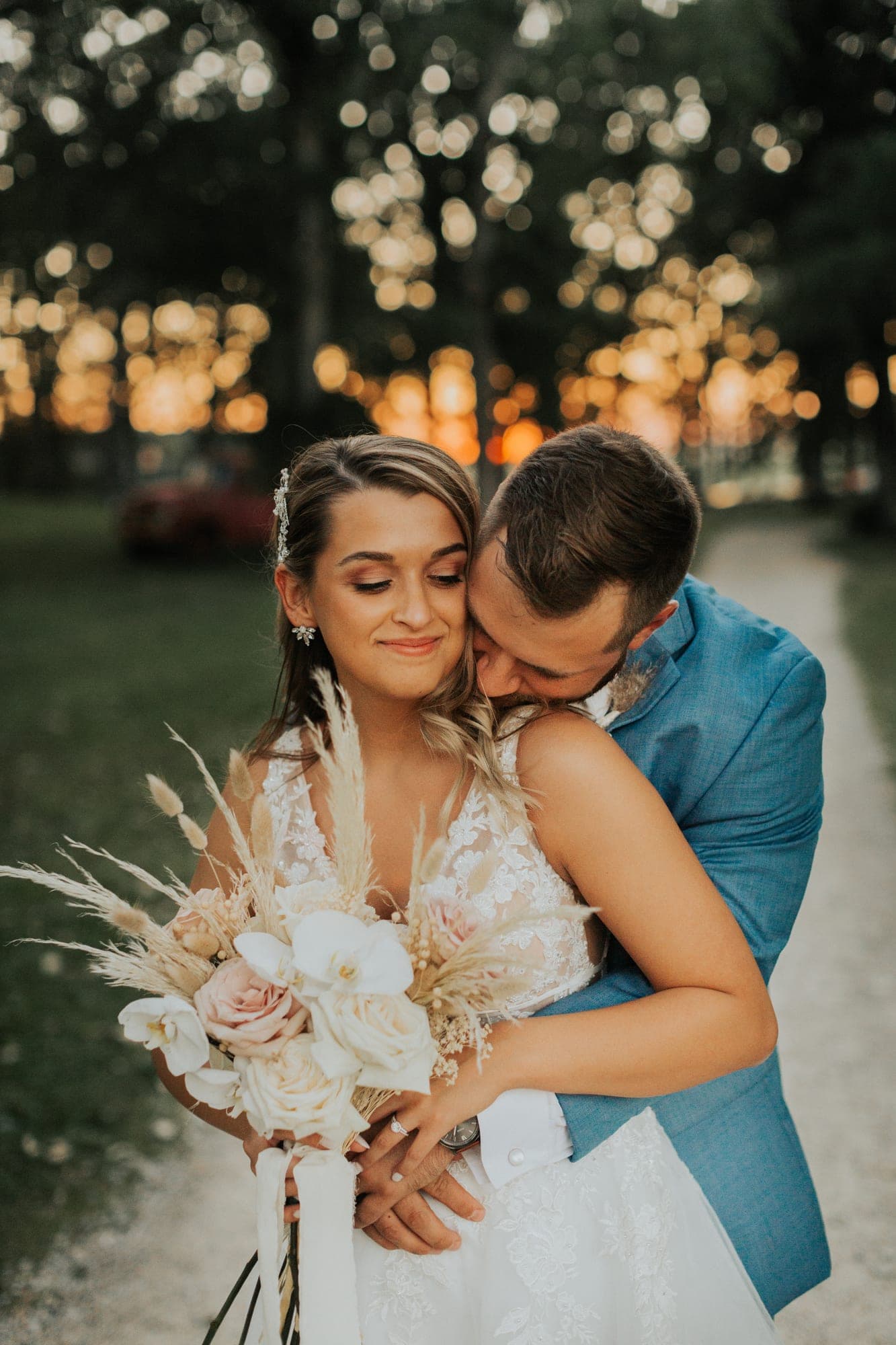 A bride and groom hold each other on a tree-lined path at dusk, string lights glowing behind them