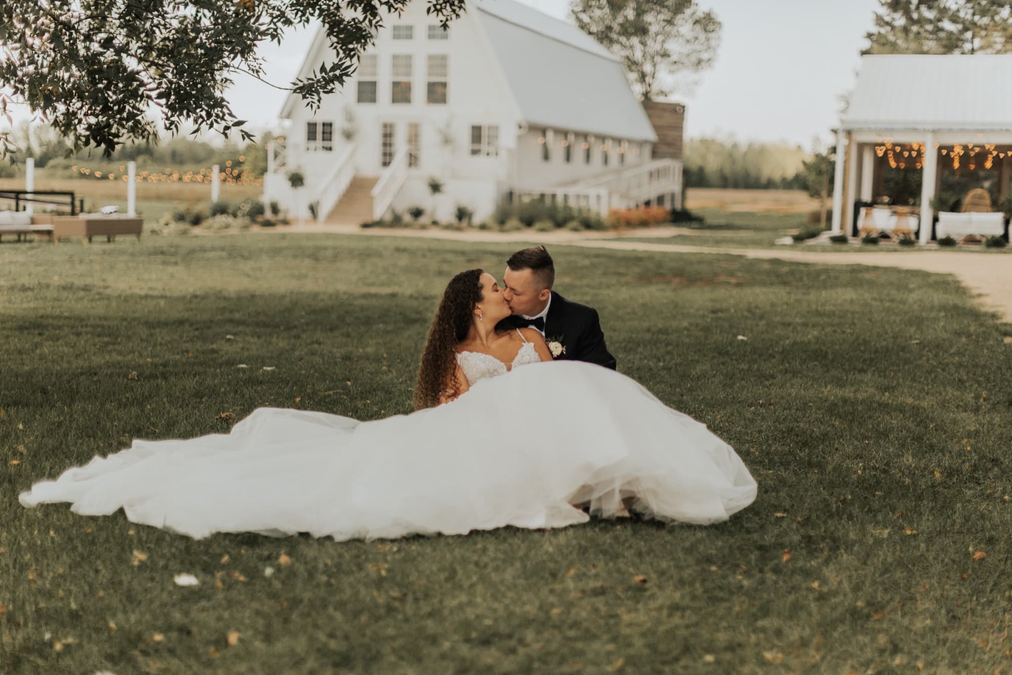 A bride and groom kiss on the lawn in front of a white barn venue, her gown spread across the grass