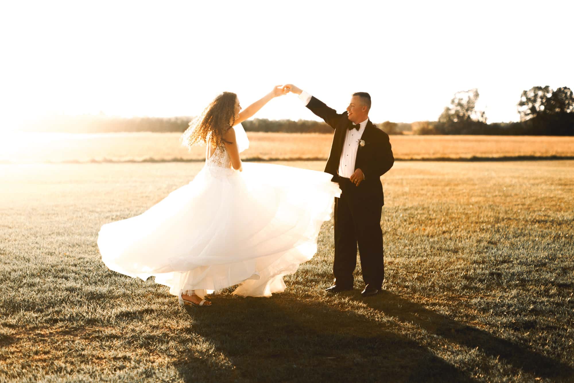 A bride in a flowing gown spins in golden evening light as her new husband holds her hand