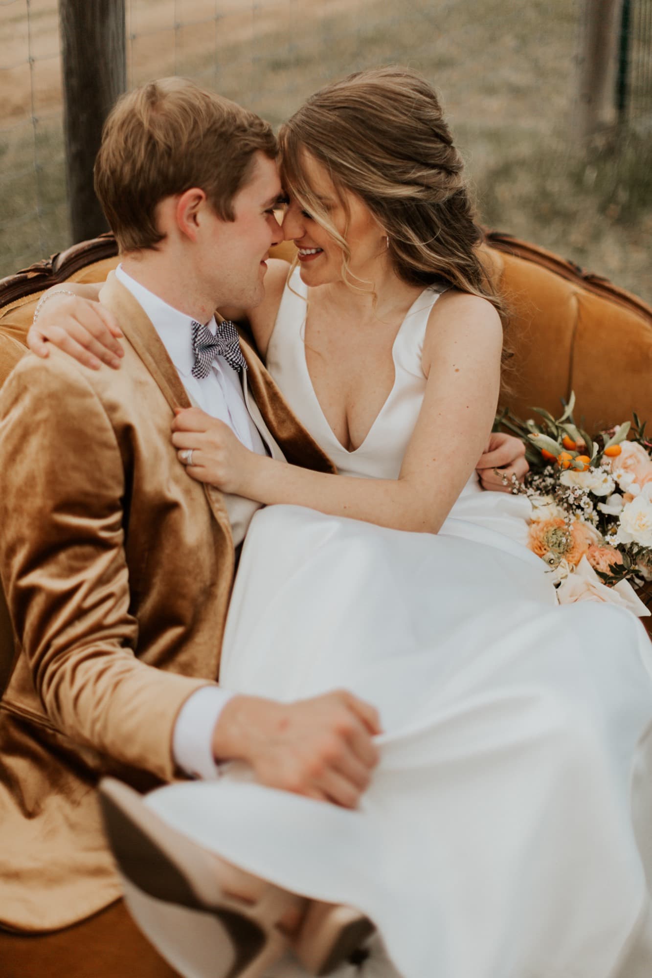 A couple sit close on a velvet couch, the groom in a cognac jacket, their foreheads nearly touching