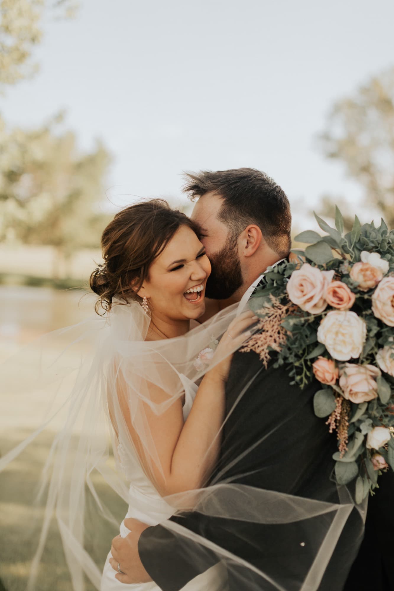 A bride laughs as her groom kisses her cheek, her veil and a bouquet of roses framing them