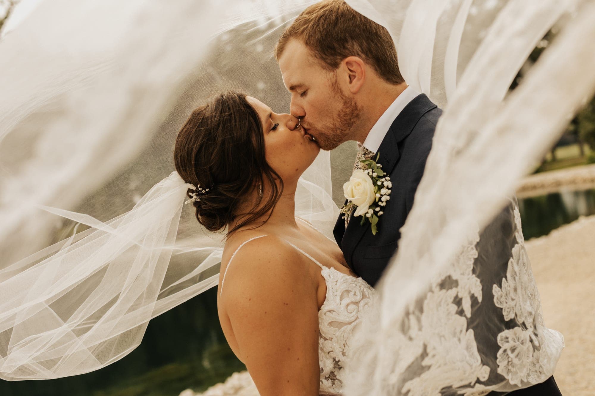 A bride and groom kiss beneath a veil lifting in the wind beside a pond