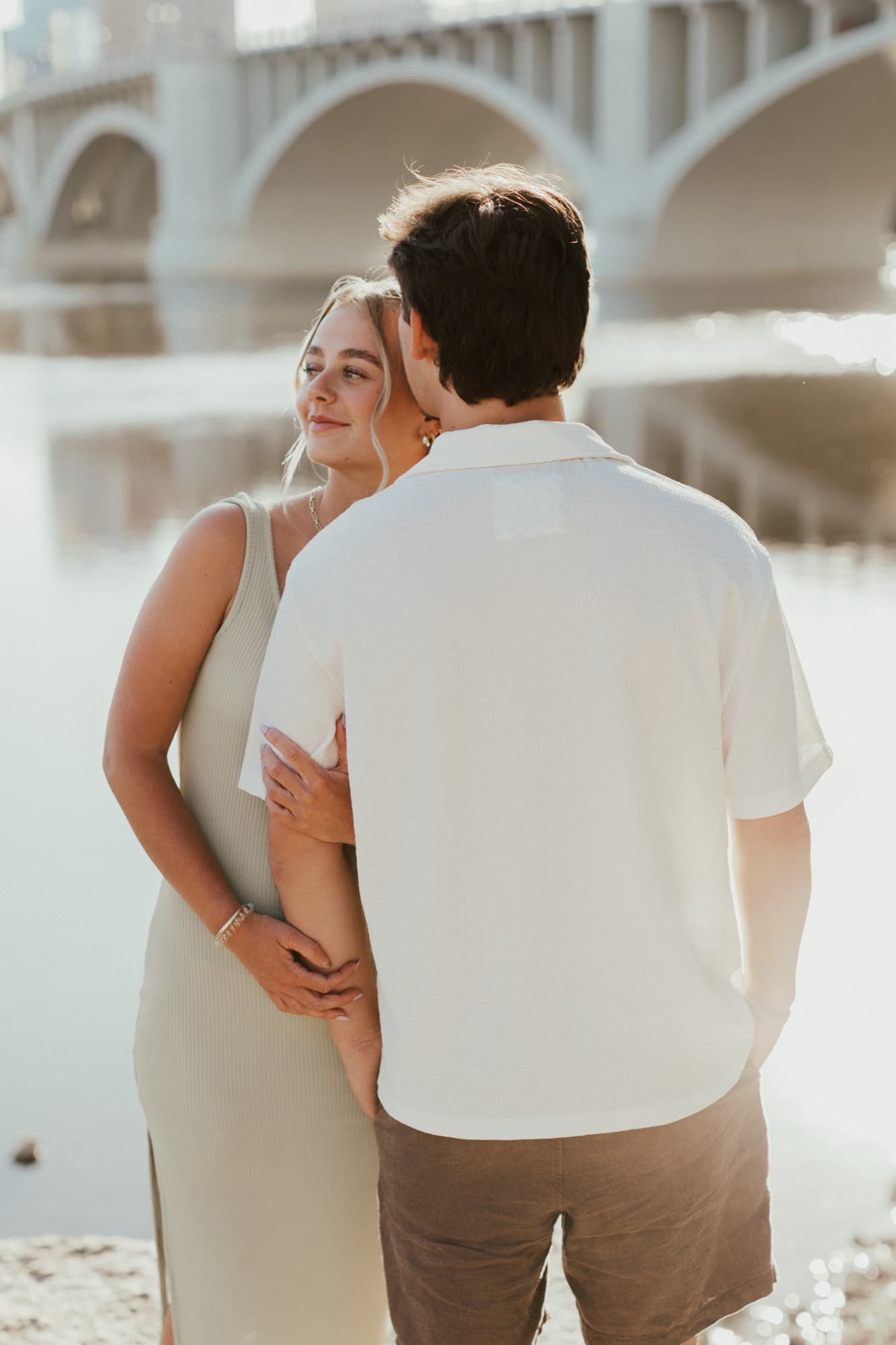 An engaged couple embrace by the Mississippi River with a Minneapolis bridge behind them in warm light