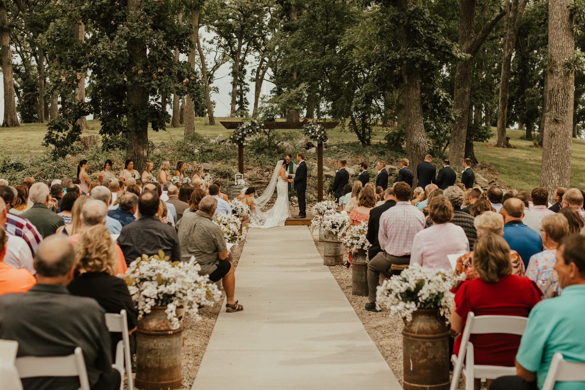 A bride and groom hold hands at a wooden floral arch as seated guests watch down a long aisle lined with milk-can flowers