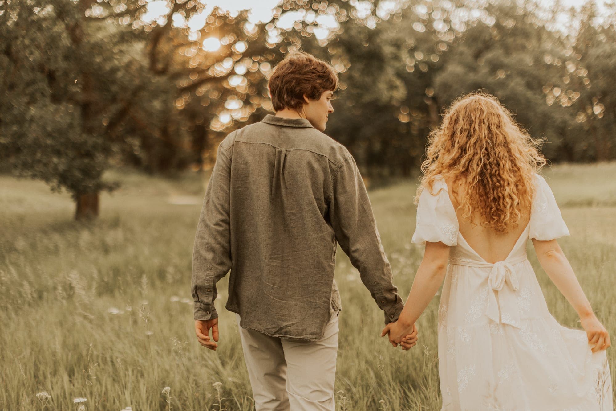 A couple walk away hand in hand through a tall grass meadow at sunset, she in a white dress and he in an olive linen shirt