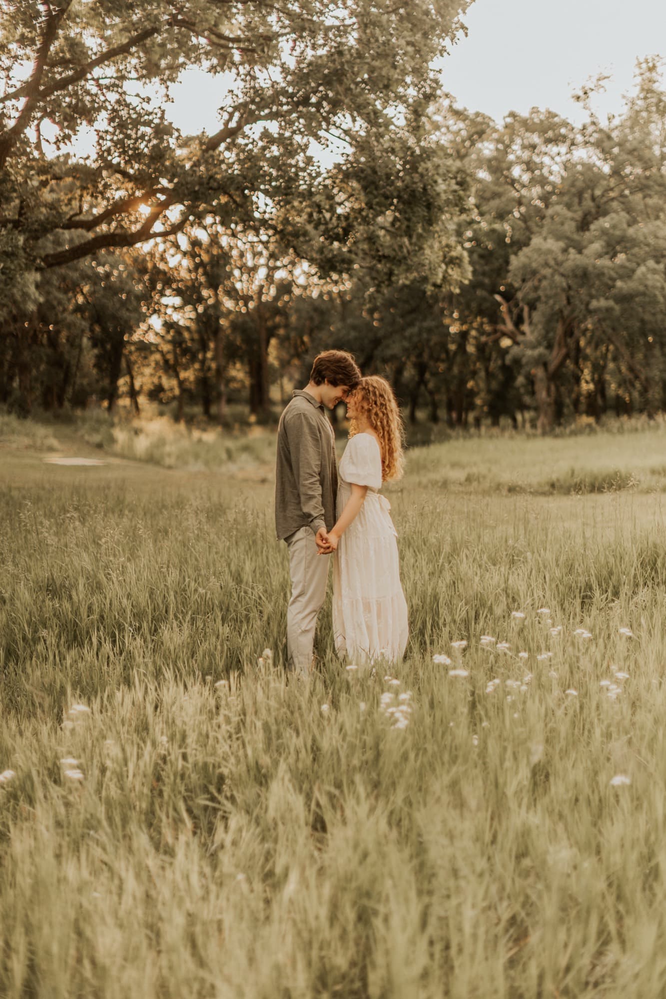 An engaged couple stand close in a sunlit meadow ringed by oak trees