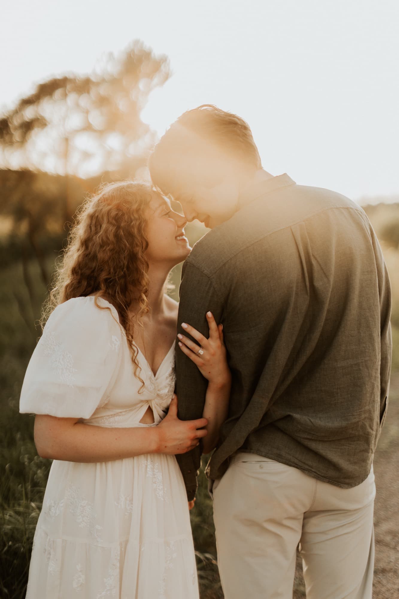 An engaged couple touch foreheads in a field, her engagement ring catching the setting sun