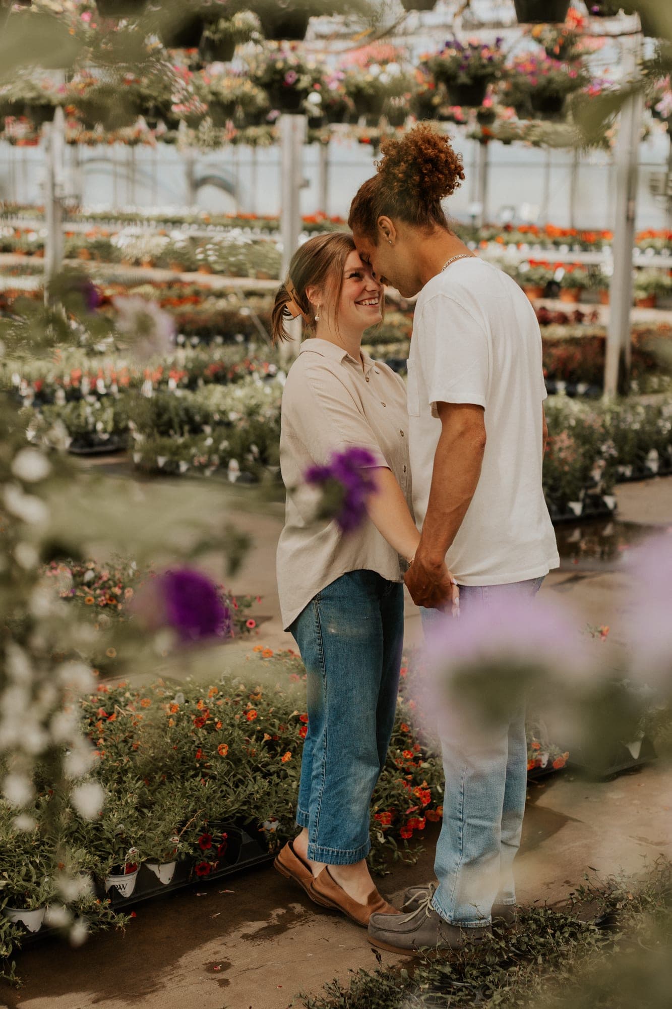 A couple hold hands and touch foreheads inside a greenhouse, framed by hanging baskets and rows of pink and purple flowers