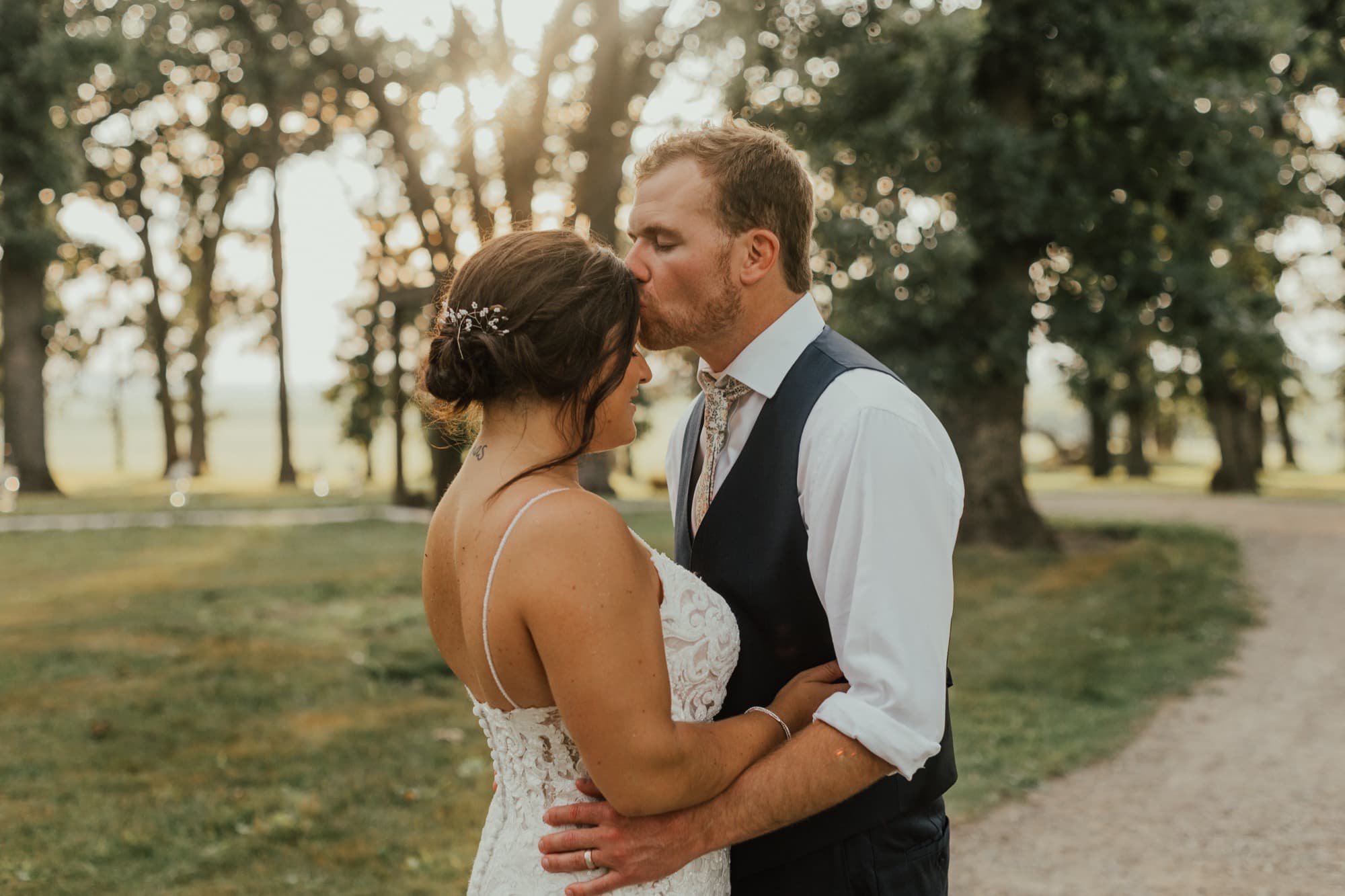 A groom kisses his bride's forehead on a tree-lined gravel path at golden hour