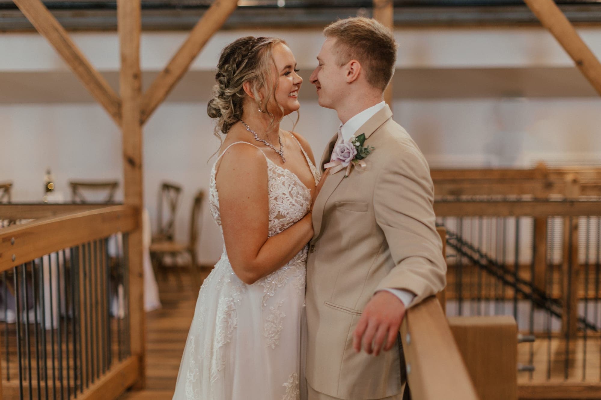 A bride in a lace gown and a groom in a tan suit lean in close and smile at each other on a wooden loft above a reception space