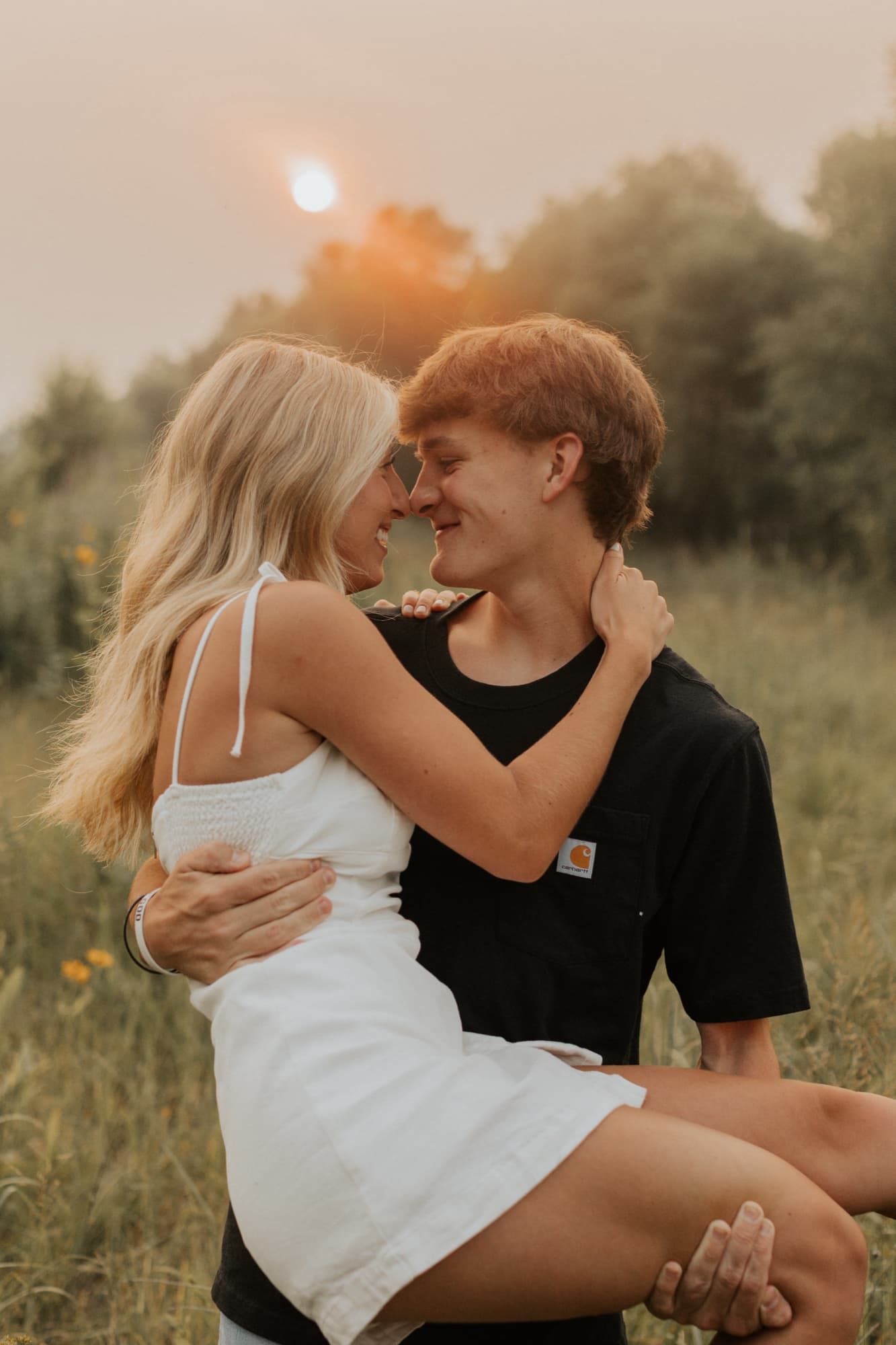 A man carries his fiancée in his arms in a meadow at hazy sunset, their noses touching as they smile