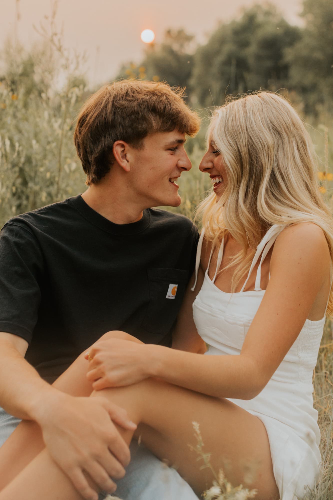 An engaged couple sit together in a meadow, laughing with their foreheads close