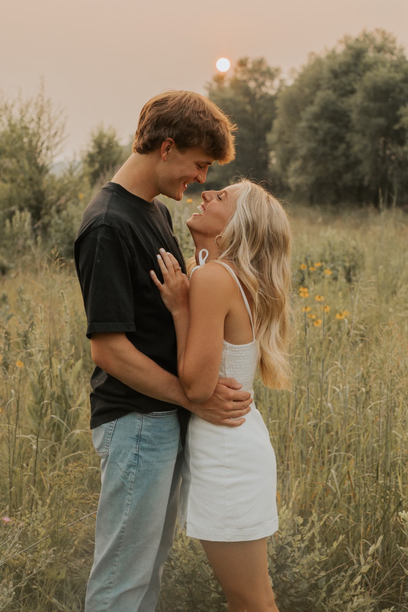 An engaged couple embrace in a field of tall grass and wildflowers at dusk