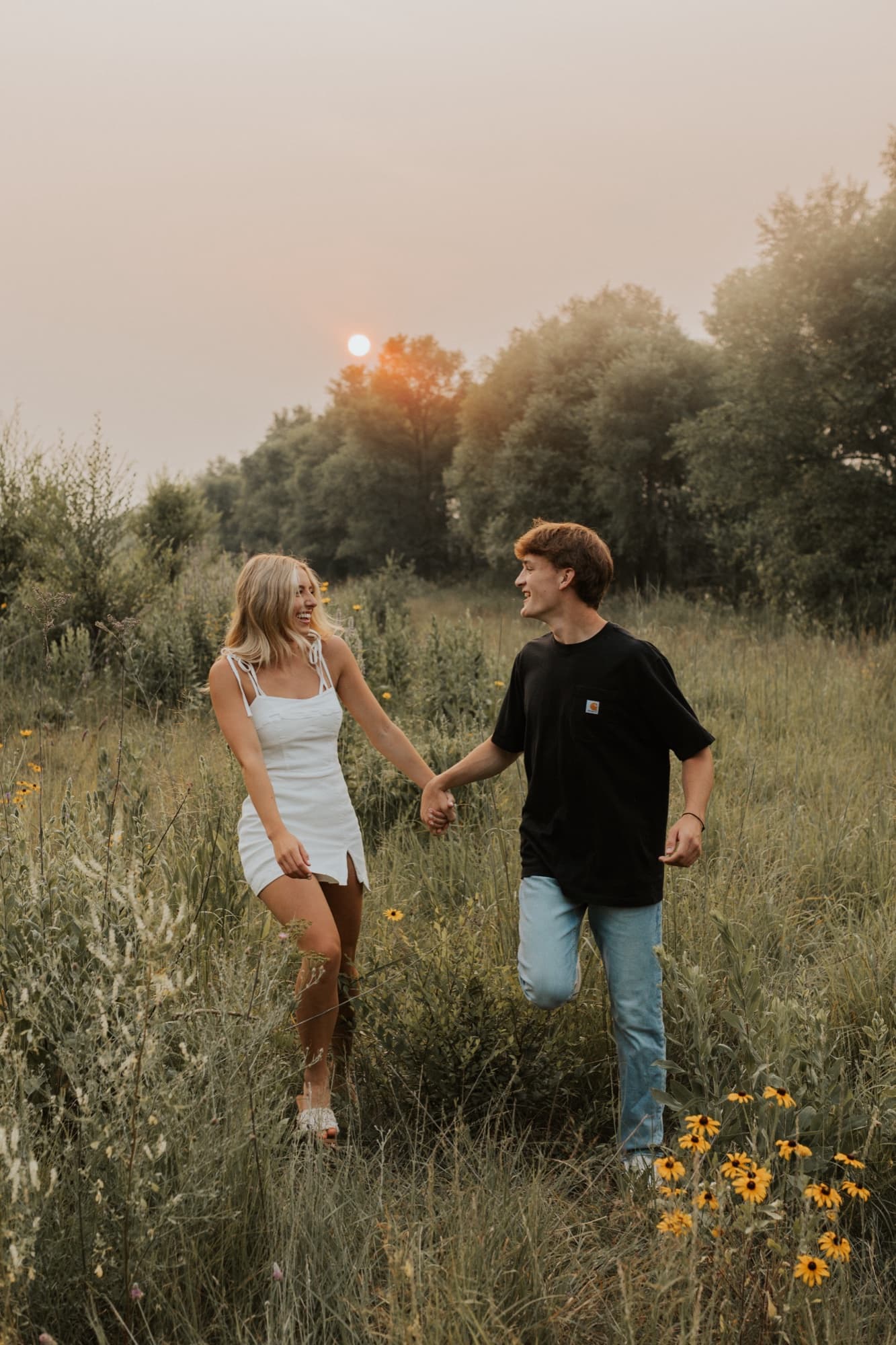 An engaged couple walk hand in hand through a meadow of black-eyed susans under a hazy sunset