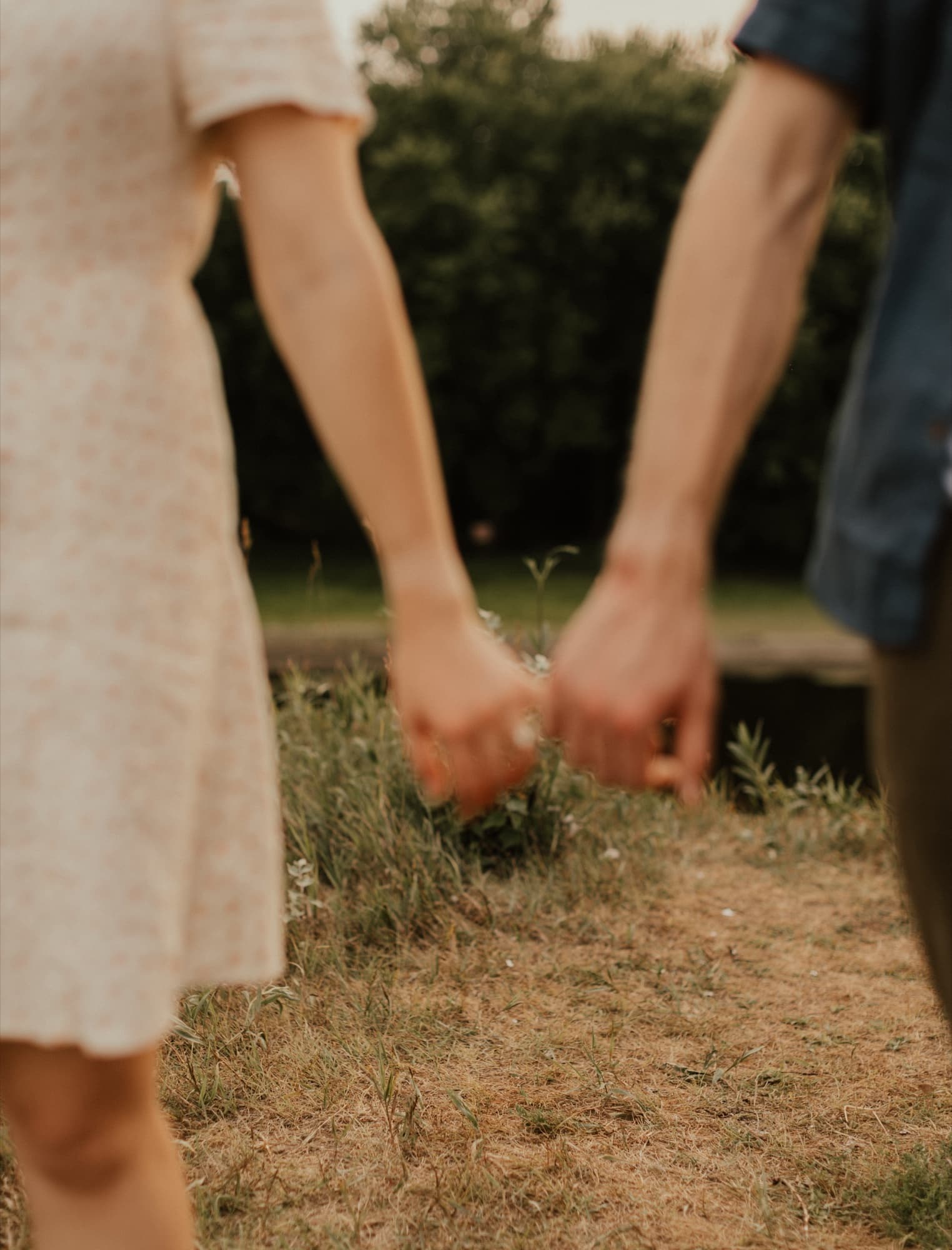 A couple's joined hands, she in a pale floral dress and he in a dark shirt, on a dry grass path