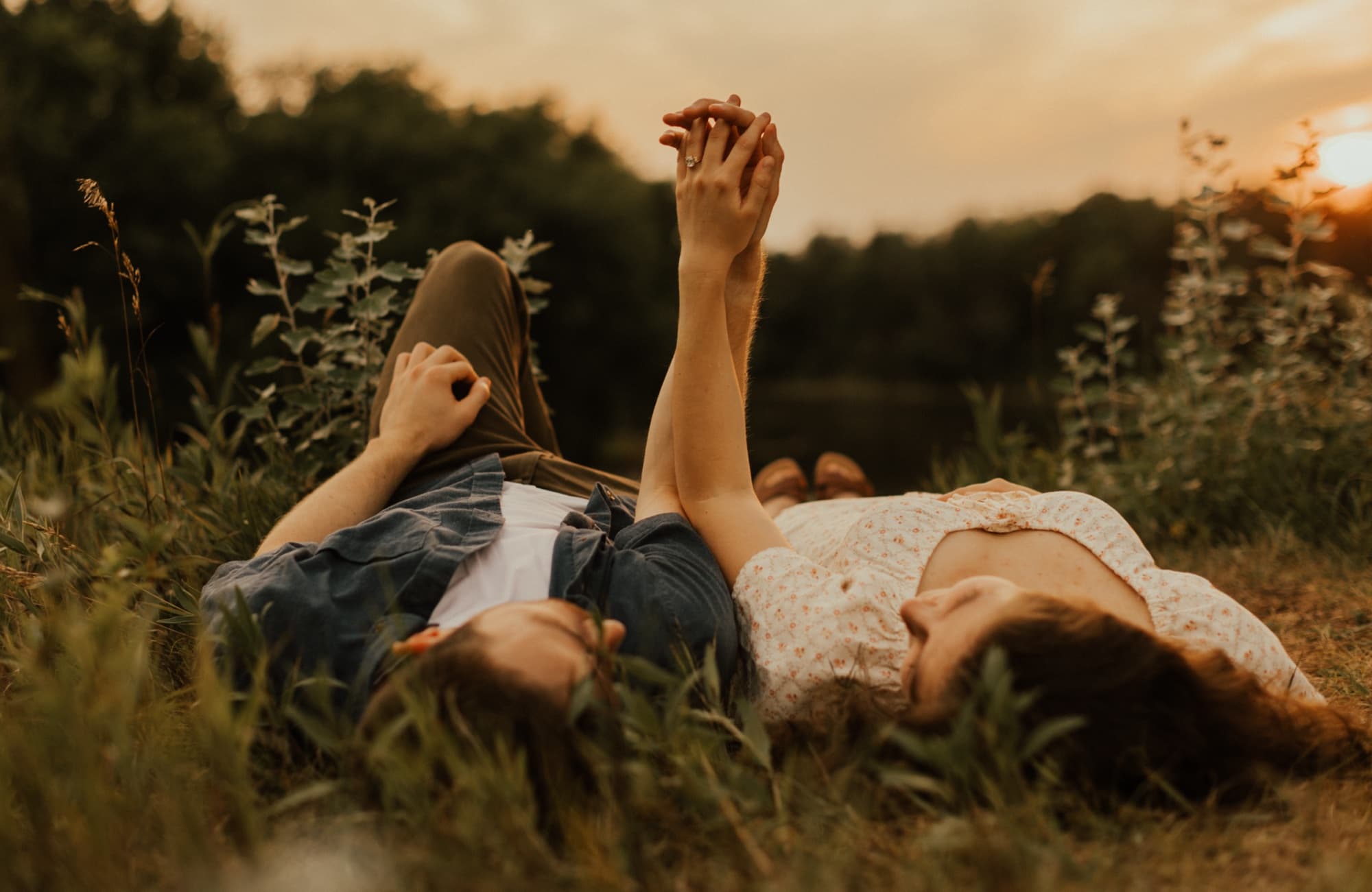 A couple lie side by side in tall grass by a pond at sunset, hands clasped and raised to the sky