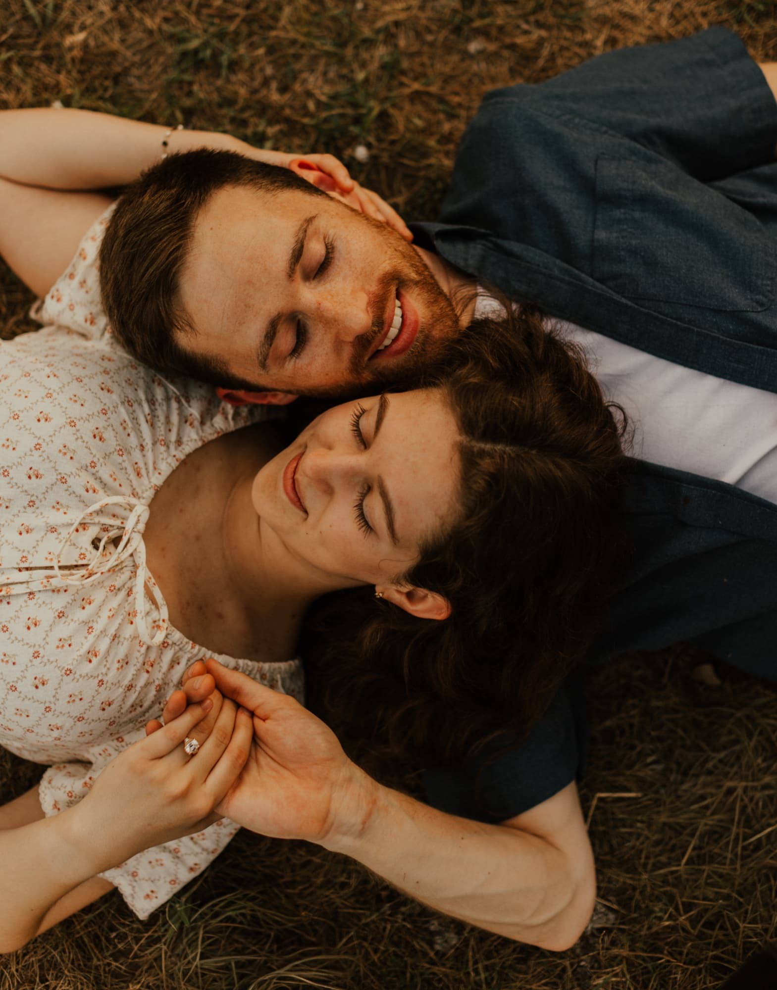 An engaged couple lie head to head in dry grass, eyes closed and smiling, her ringed hand resting in his