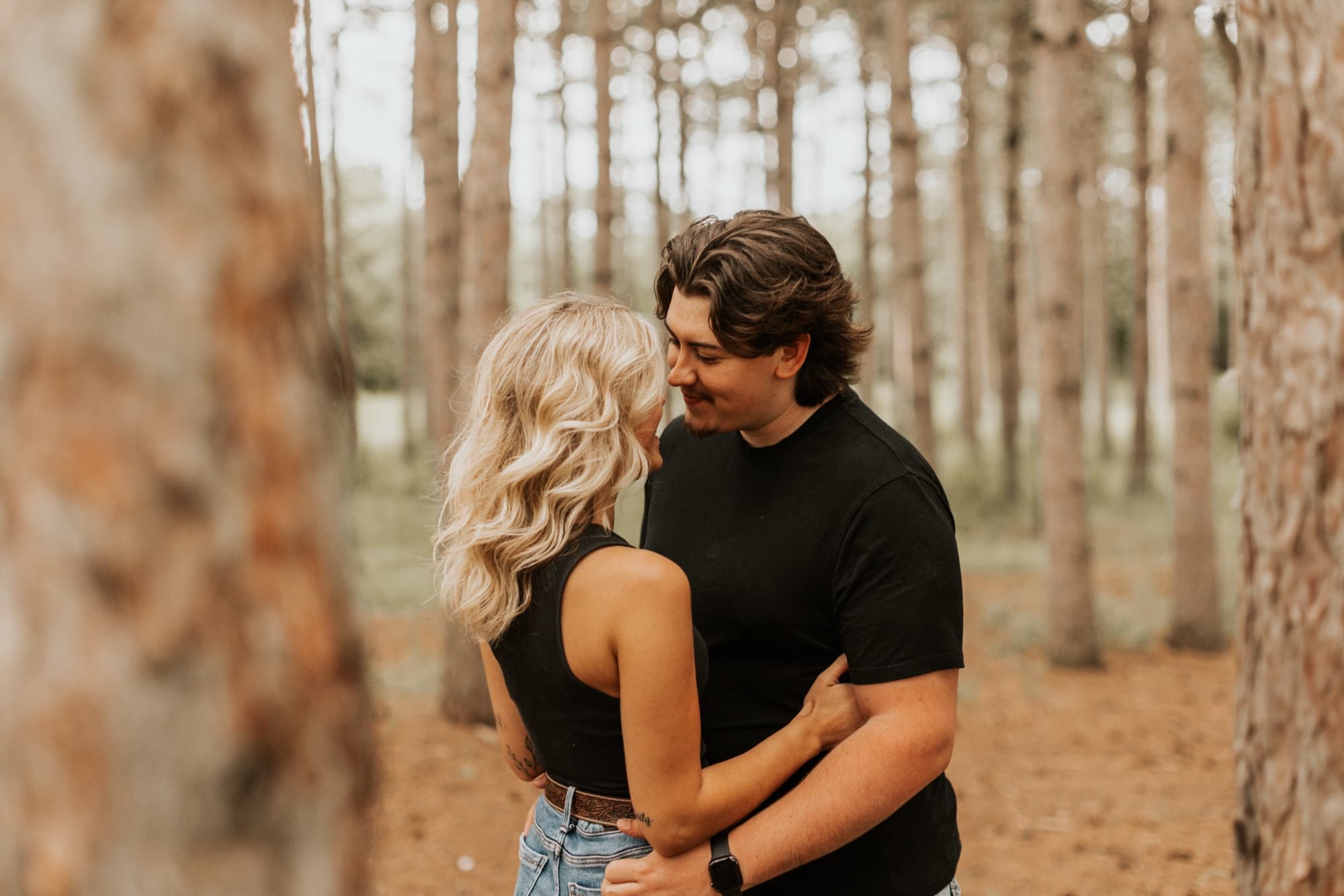 A couple embrace among tall pine trunks, smiling at each other