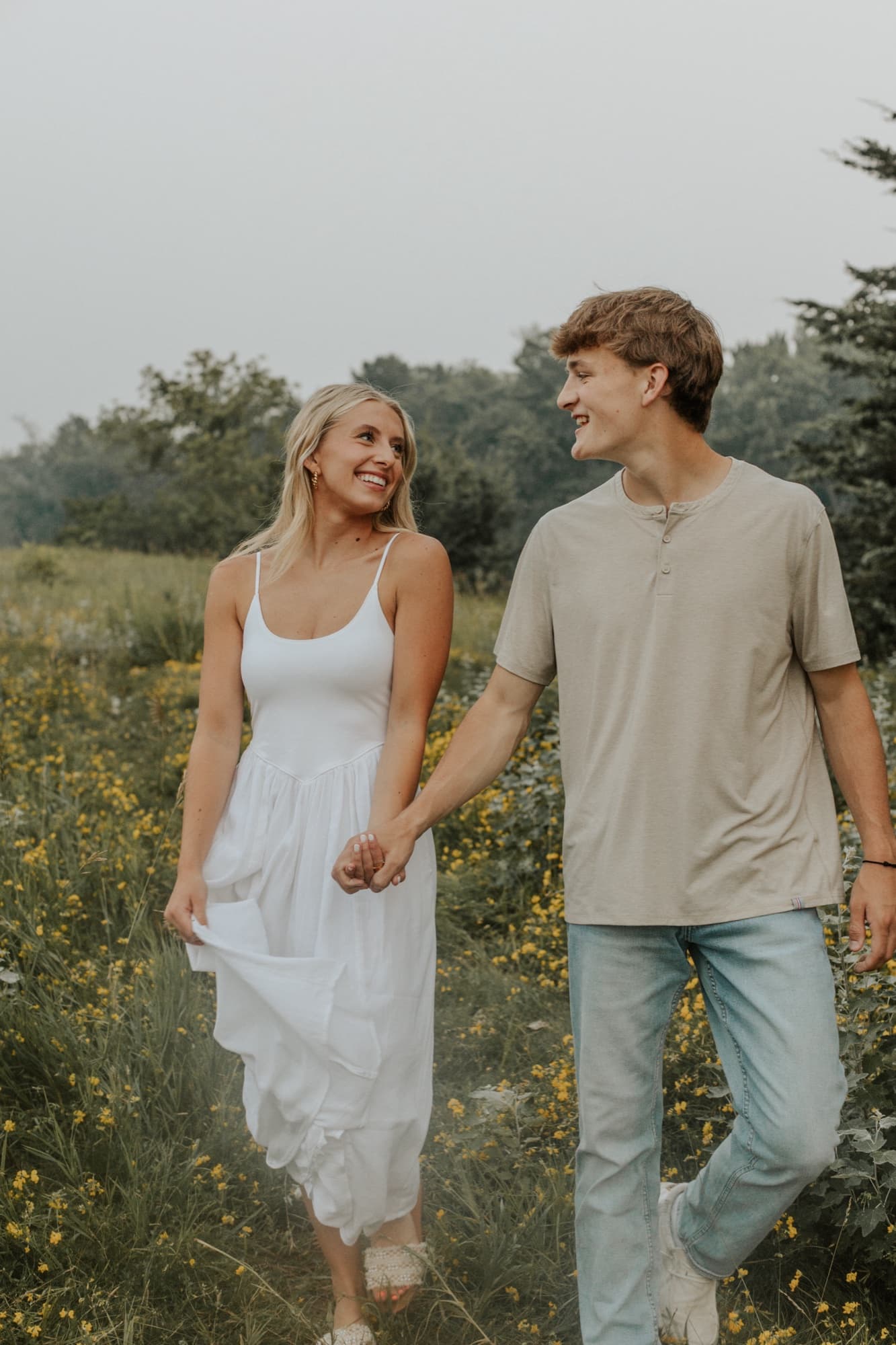 An engaged couple walk hand in hand through a meadow of yellow wildflowers, laughing at each other, she in a long white dress