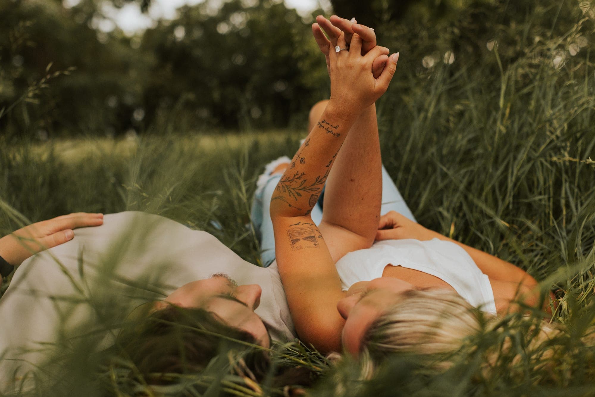 A couple lie in tall grass with their hands clasped in the air, her engagement ring catching the light