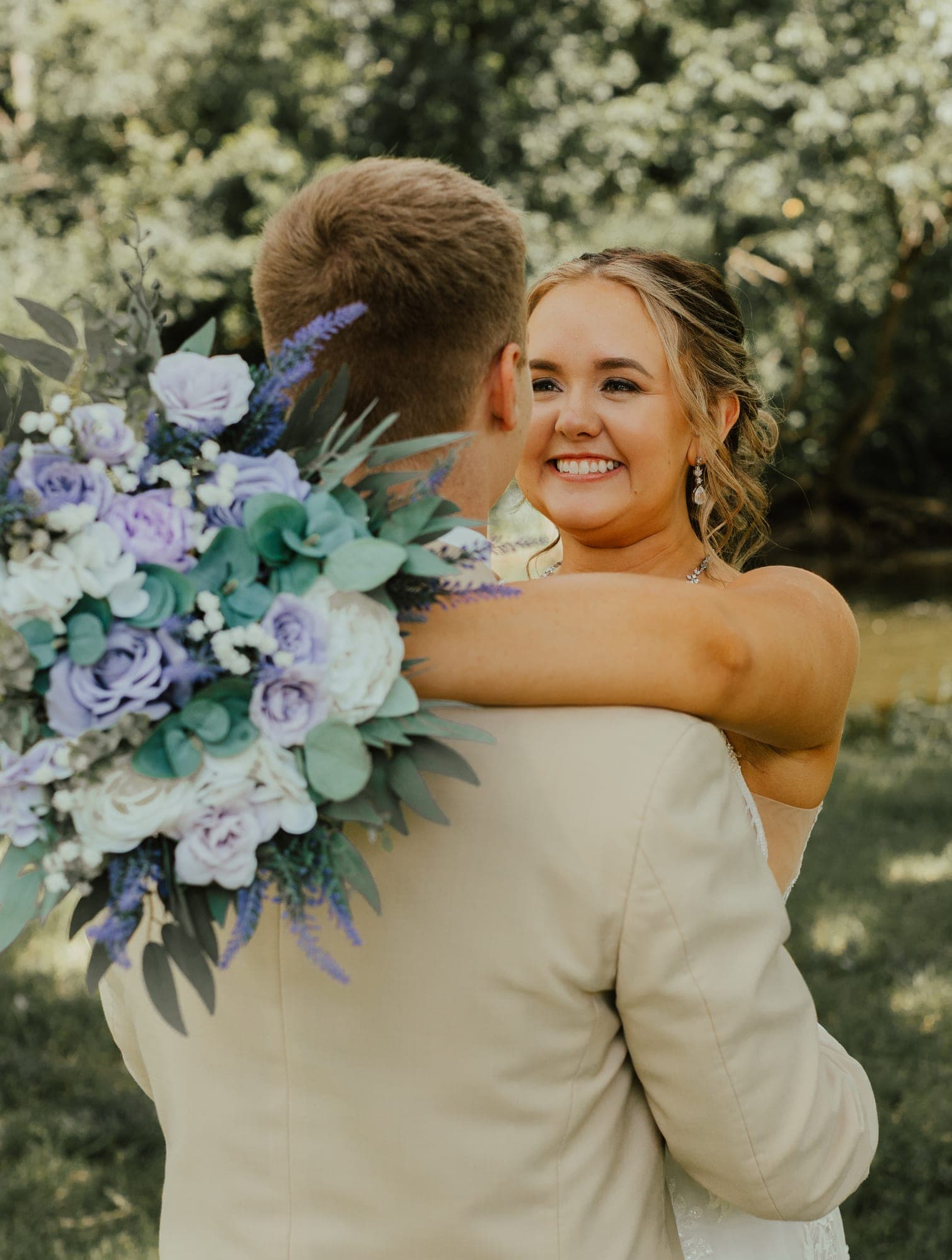 A bride wraps her arms around her groom's neck and smiles at him, holding a lavender and white bouquet over his shoulder