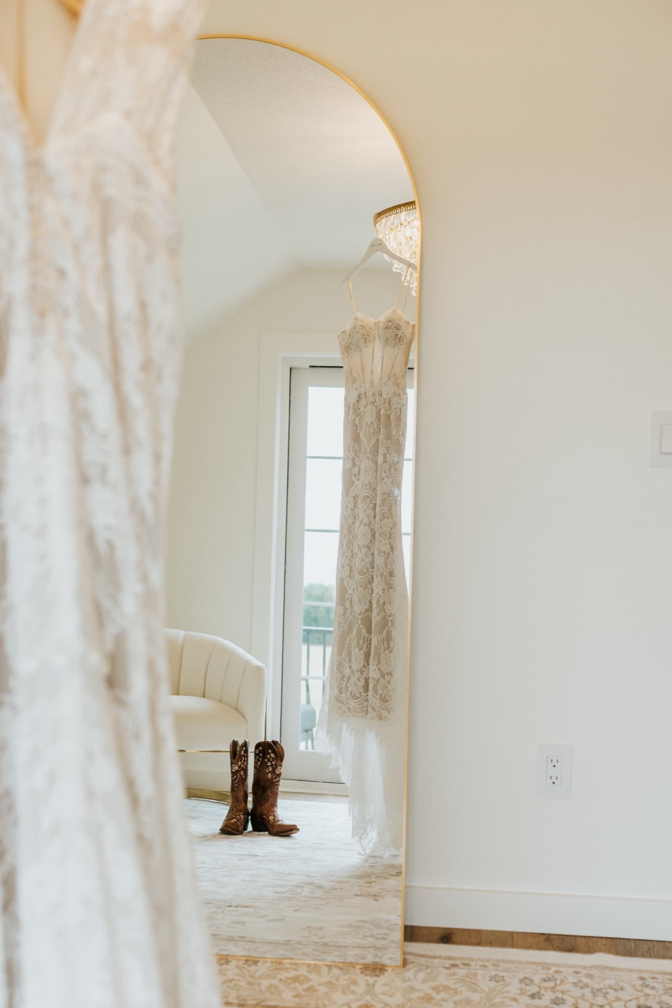 A lace wedding gown hangs in an arched gold mirror, a pair of floral-tooled cowboy boots on the floor beneath it