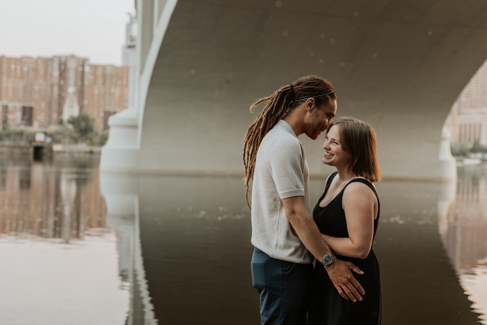 An engaged couple touch foreheads beneath a Minneapolis river bridge, the city beyond