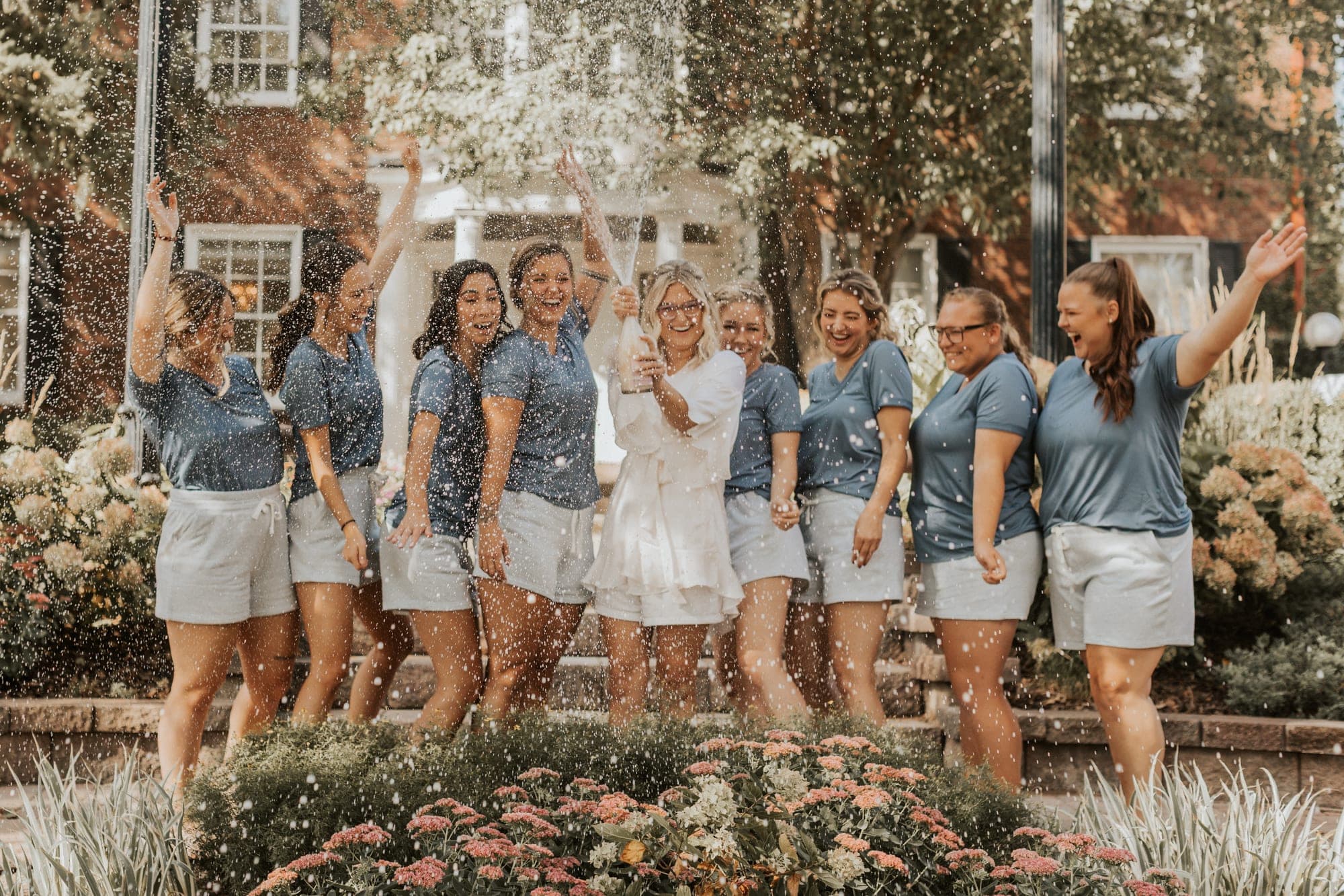 A bride in white sprays champagne over her bridesmaids in matching blue shirts as they cheer in a garden