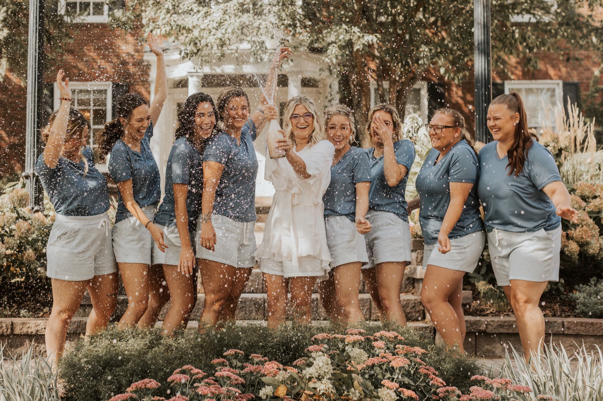 A circle of women in matching outfits toss confetti, the bride laughing in white at the center