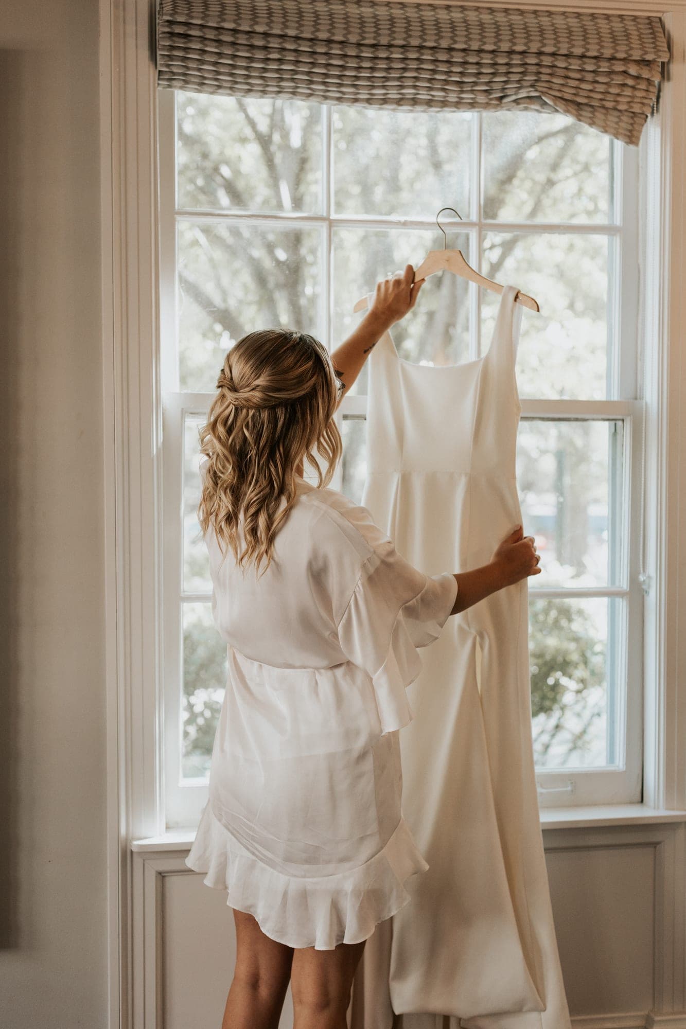 A woman in a white robe holds up a wedding dress on a wooden hanger against a bright window with trees outside