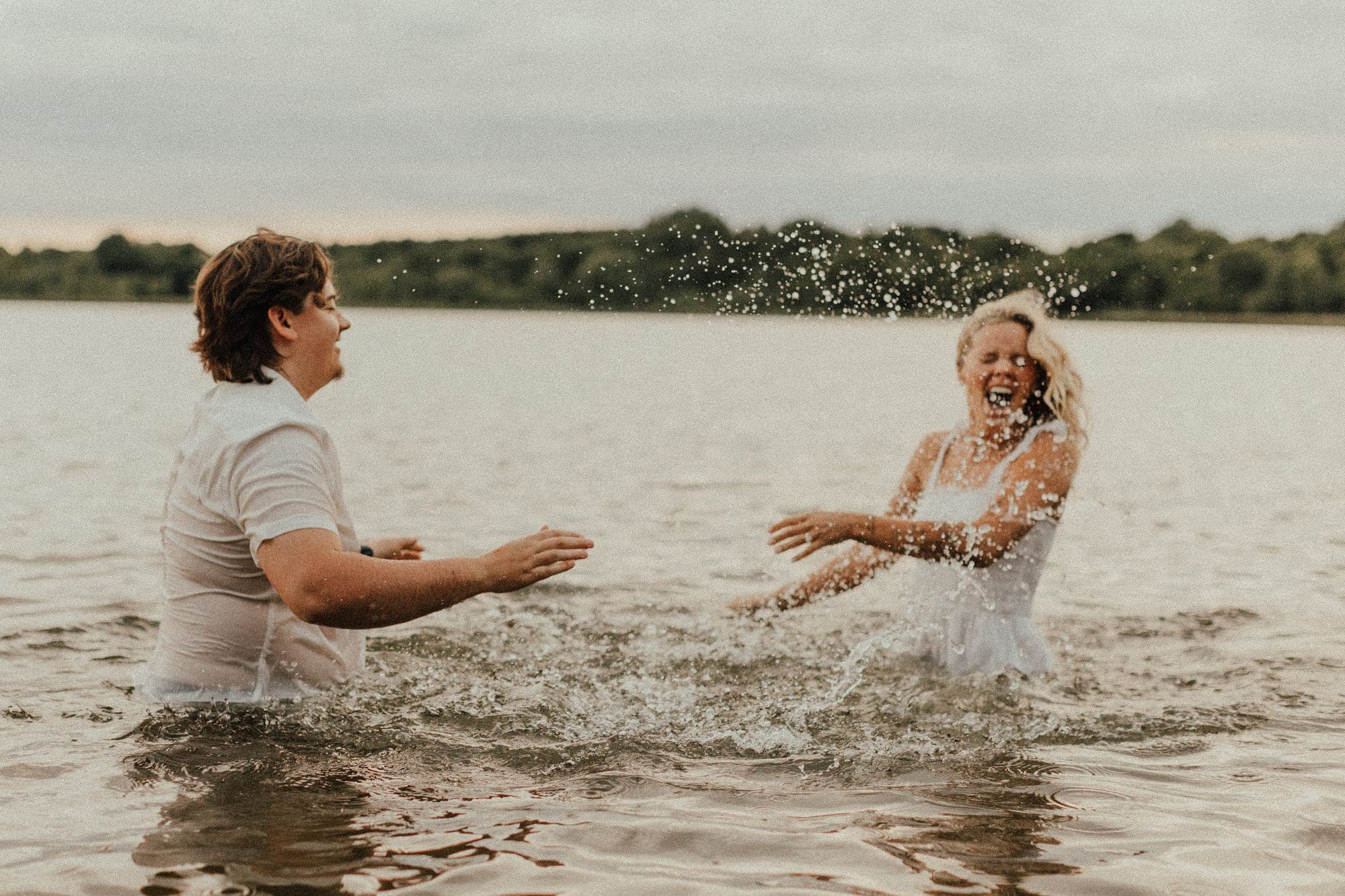 A couple in white splash water at each other in a lake at dusk, the woman laughing
