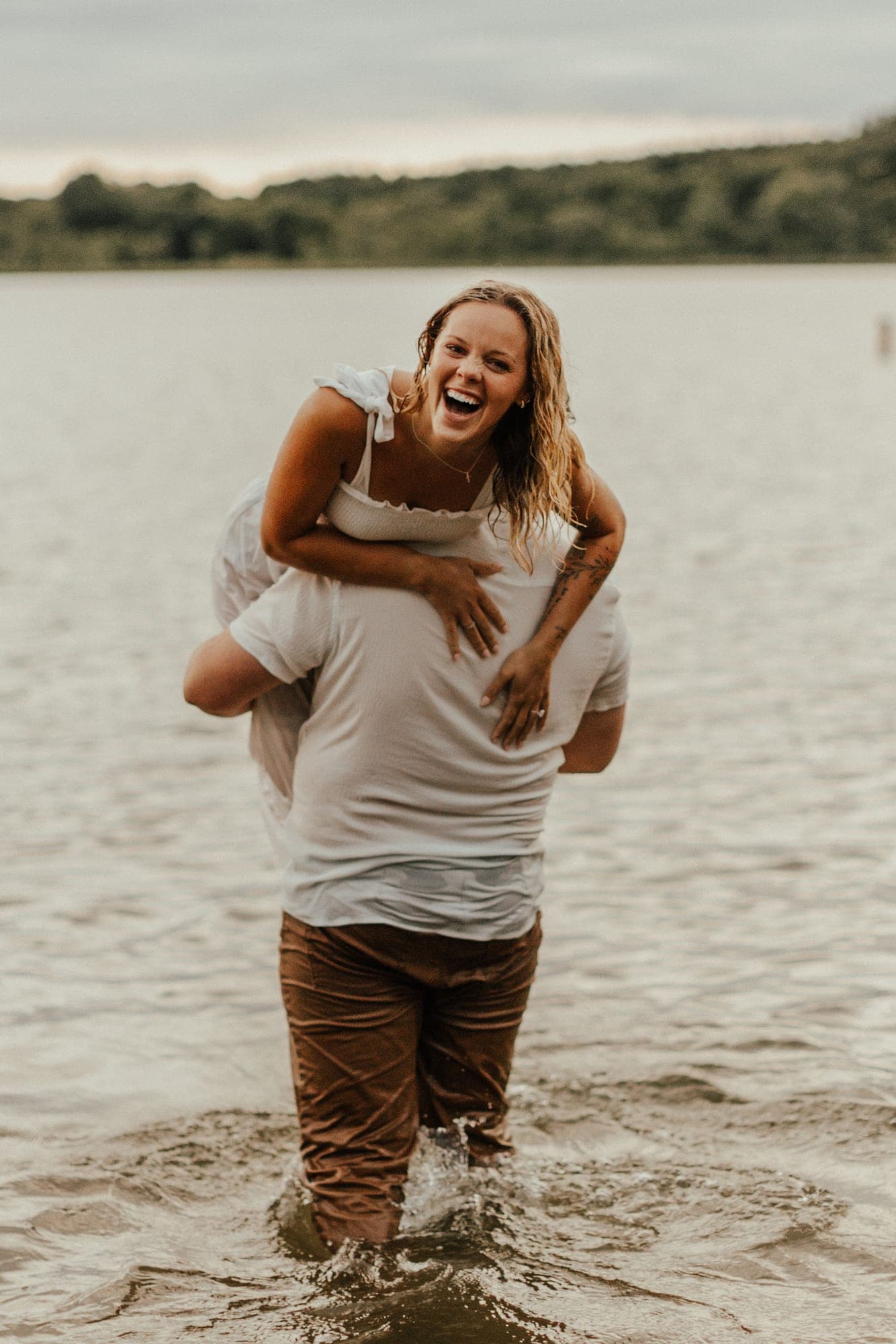A woman rides piggyback on a man wading through a lake, both soaked and laughing toward the camera