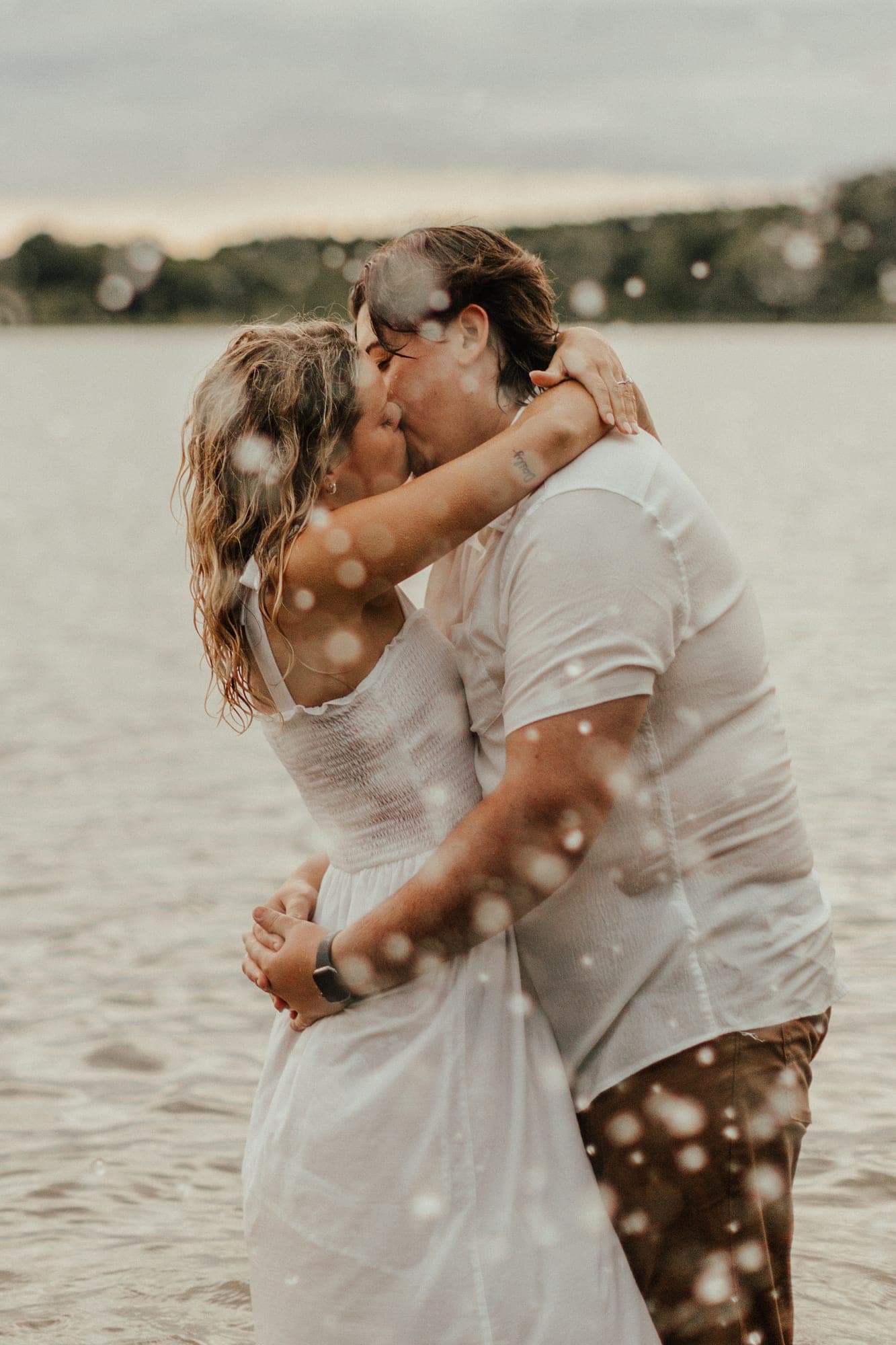 A couple in white kiss while standing waist-deep in a lake at dusk, water droplets suspended around them