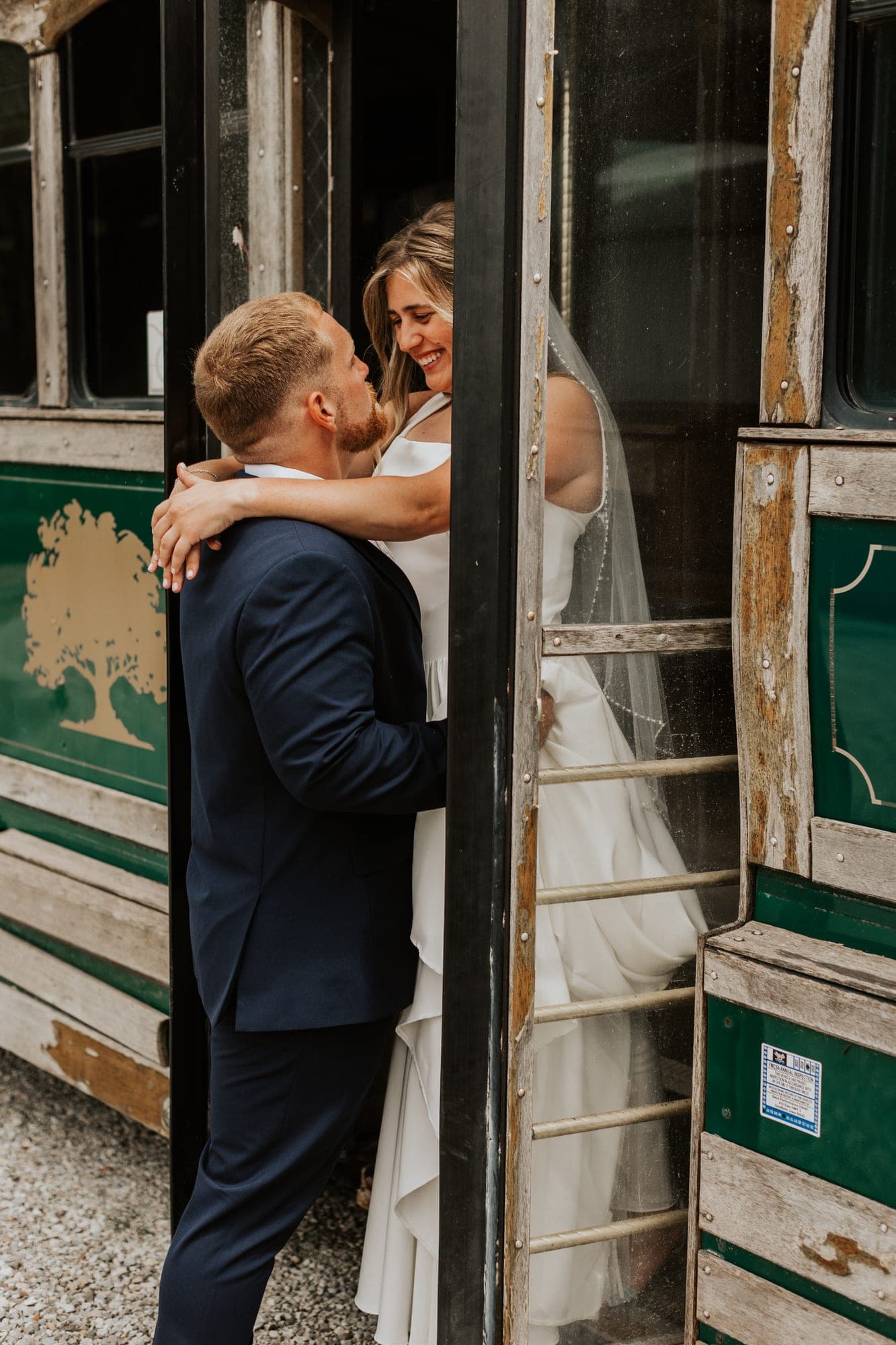 A bride in a veil wraps her arms around a groom in a navy suit, both smiling, framed in the open door of a green trolley