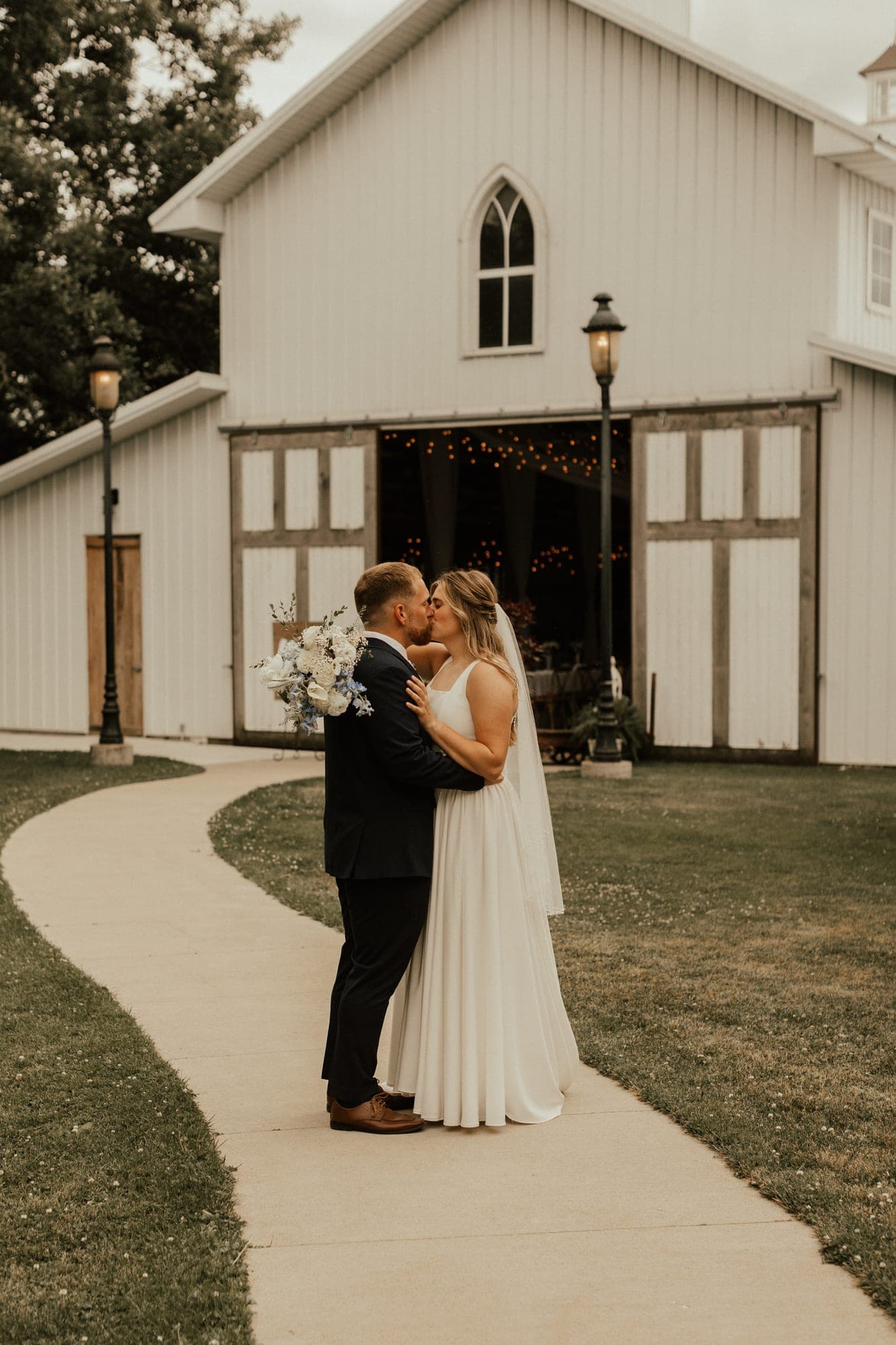 A bride and groom kiss on a curved path in front of a white barn venue, she in a long veil holding white and pale-blue flowers