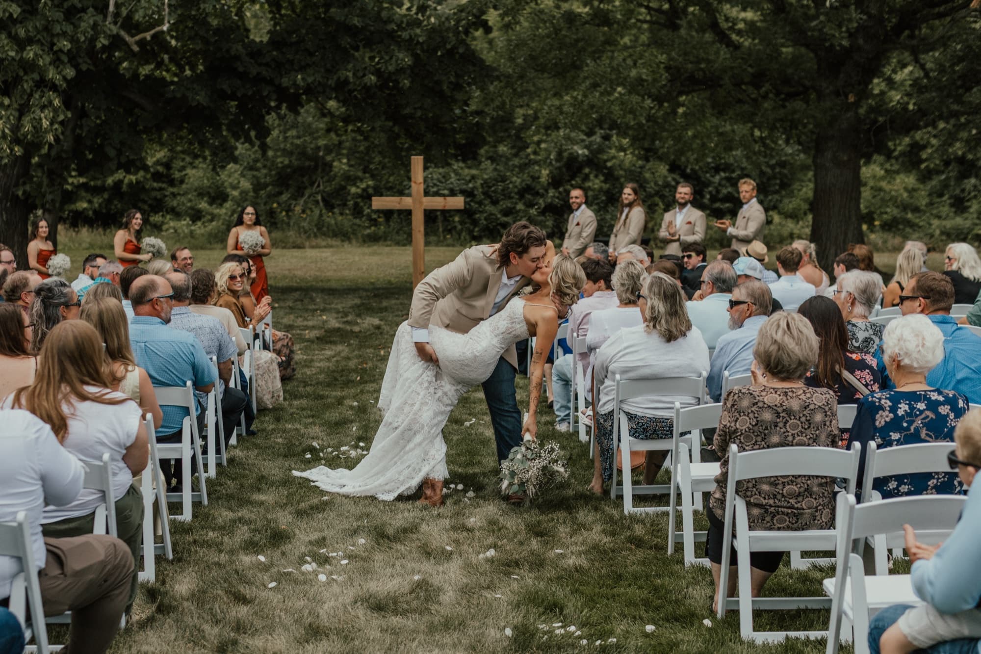 Betsy and Caleb share a kiss at their outdoor ceremony beneath a wooden cross, guests seated on white chairs on both sides