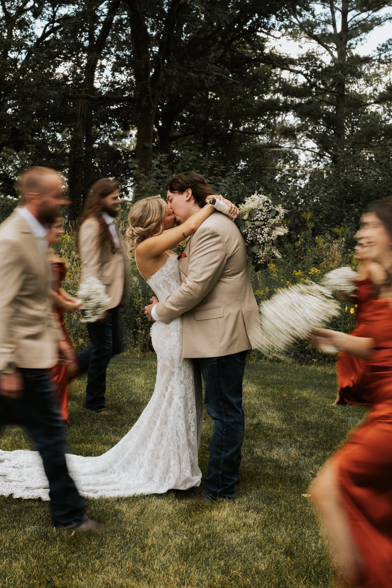 A bride and groom kiss as their wedding party in rust dresses and tan jackets moves past in a blur