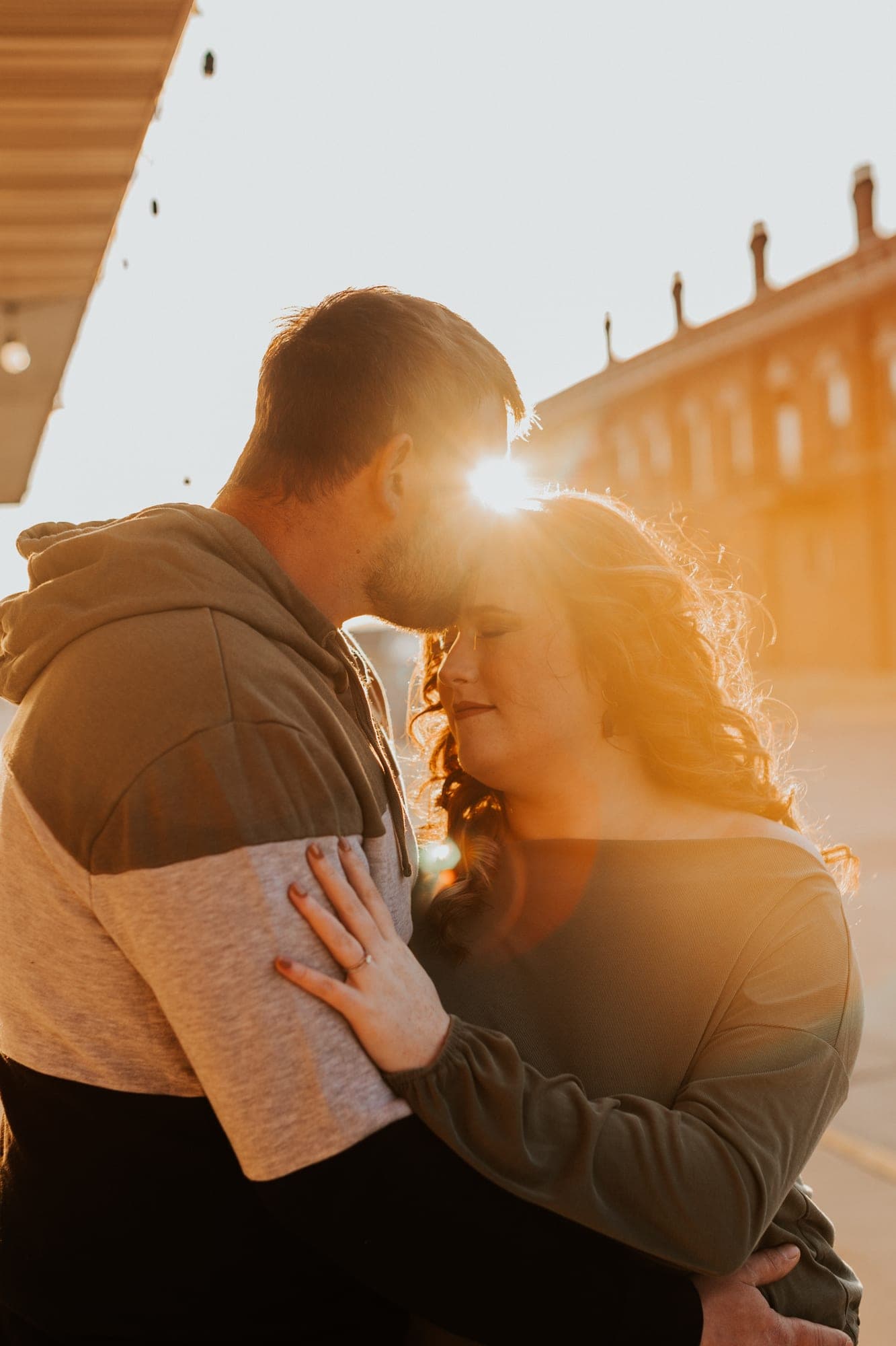 A couple lean their foreheads together in warm backlit sun, her ringed hand resting on his chest as the light flares between them