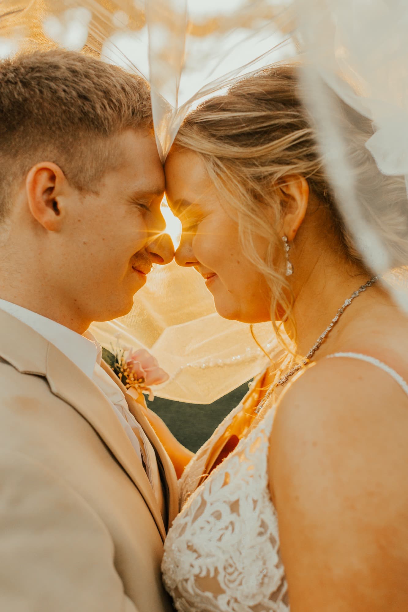 A bride and groom rest their foreheads together beneath her veil, lit by intense golden sun