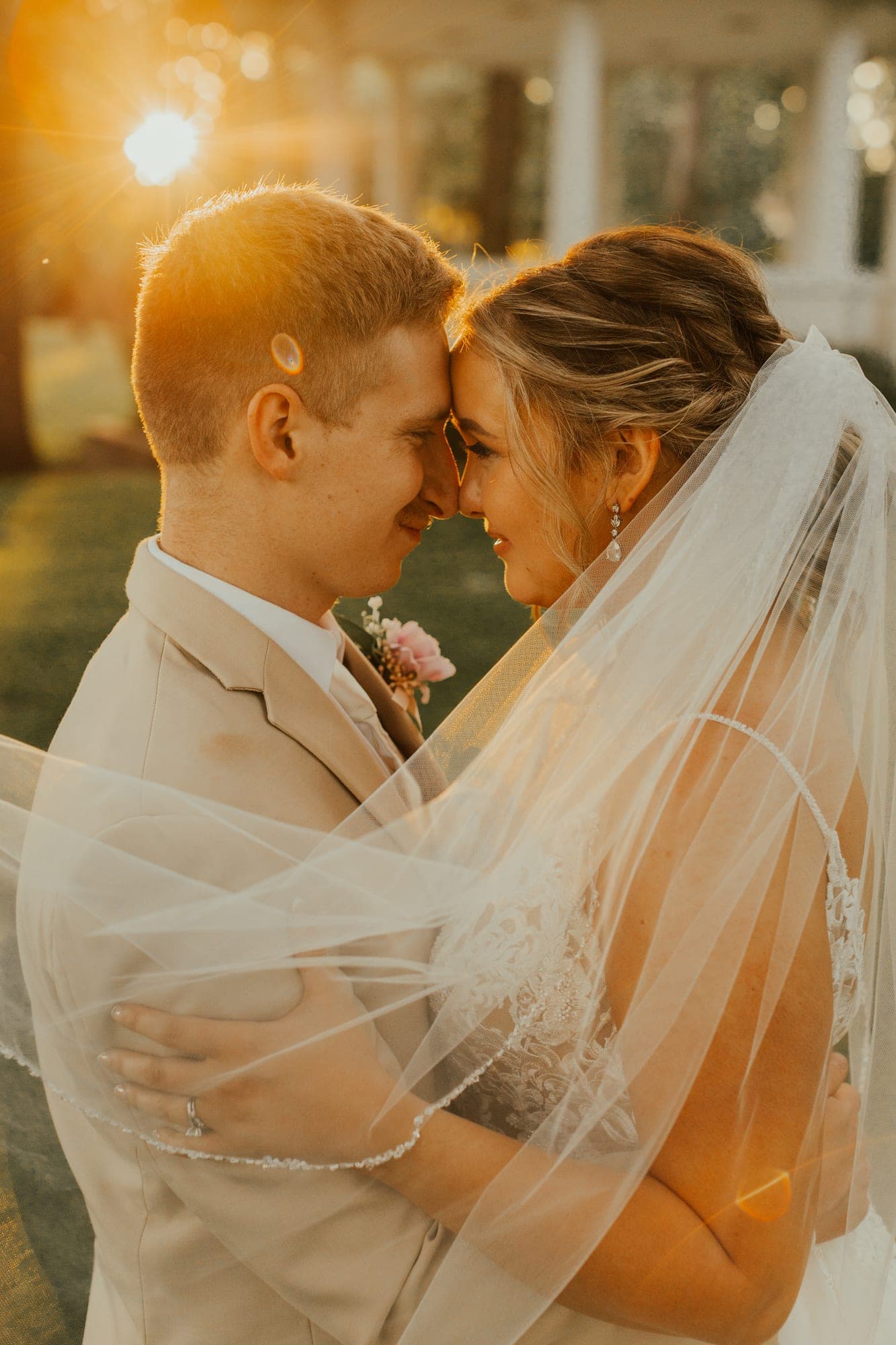 A bride and groom press their foreheads together at sunset, her veil sweeping across his suit as golden light flares behind them