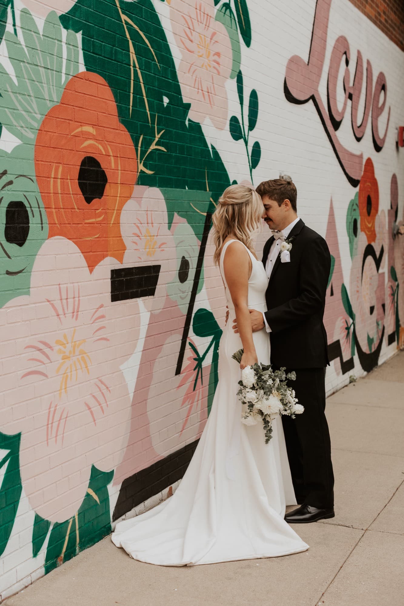 A bride and groom touch foreheads in front of a painted floral mural reading 'Love' in the city