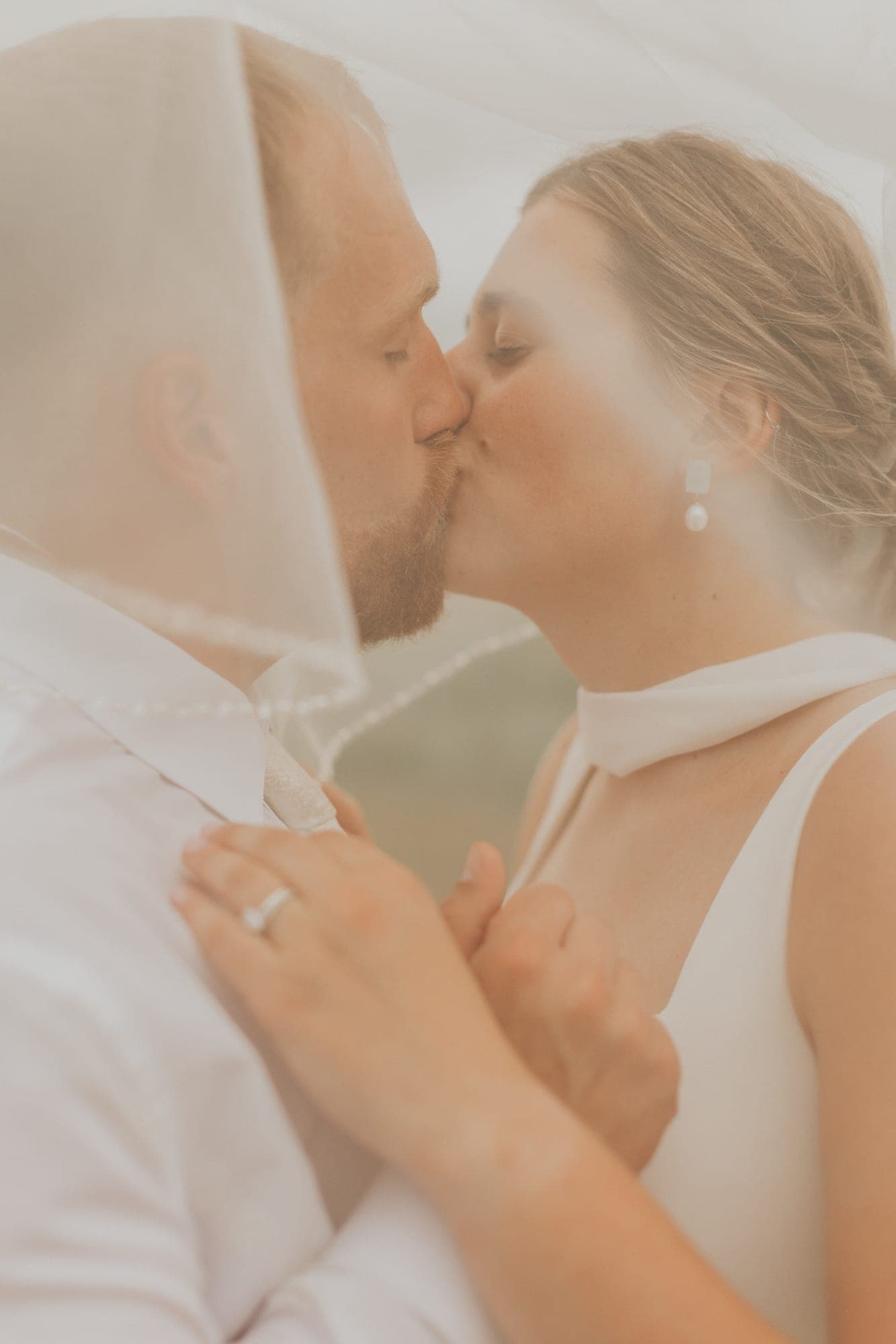 A bride and groom kiss beneath her sheer veil in soft pale light, her ring hand resting between them