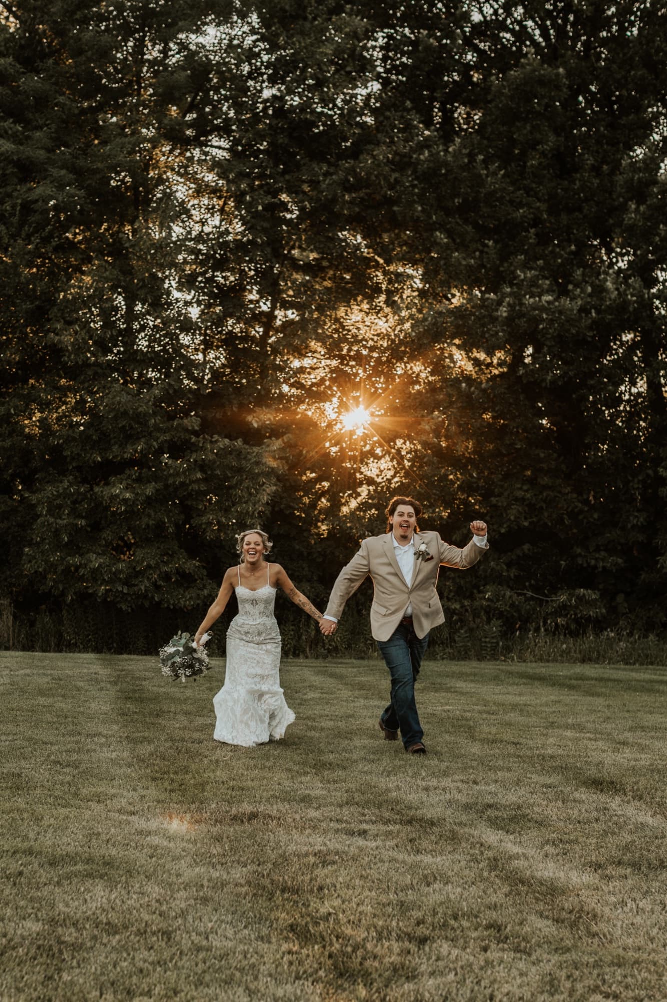 A bride and groom run hand in hand across a lawn, laughing, as the sun bursts through the trees behind them