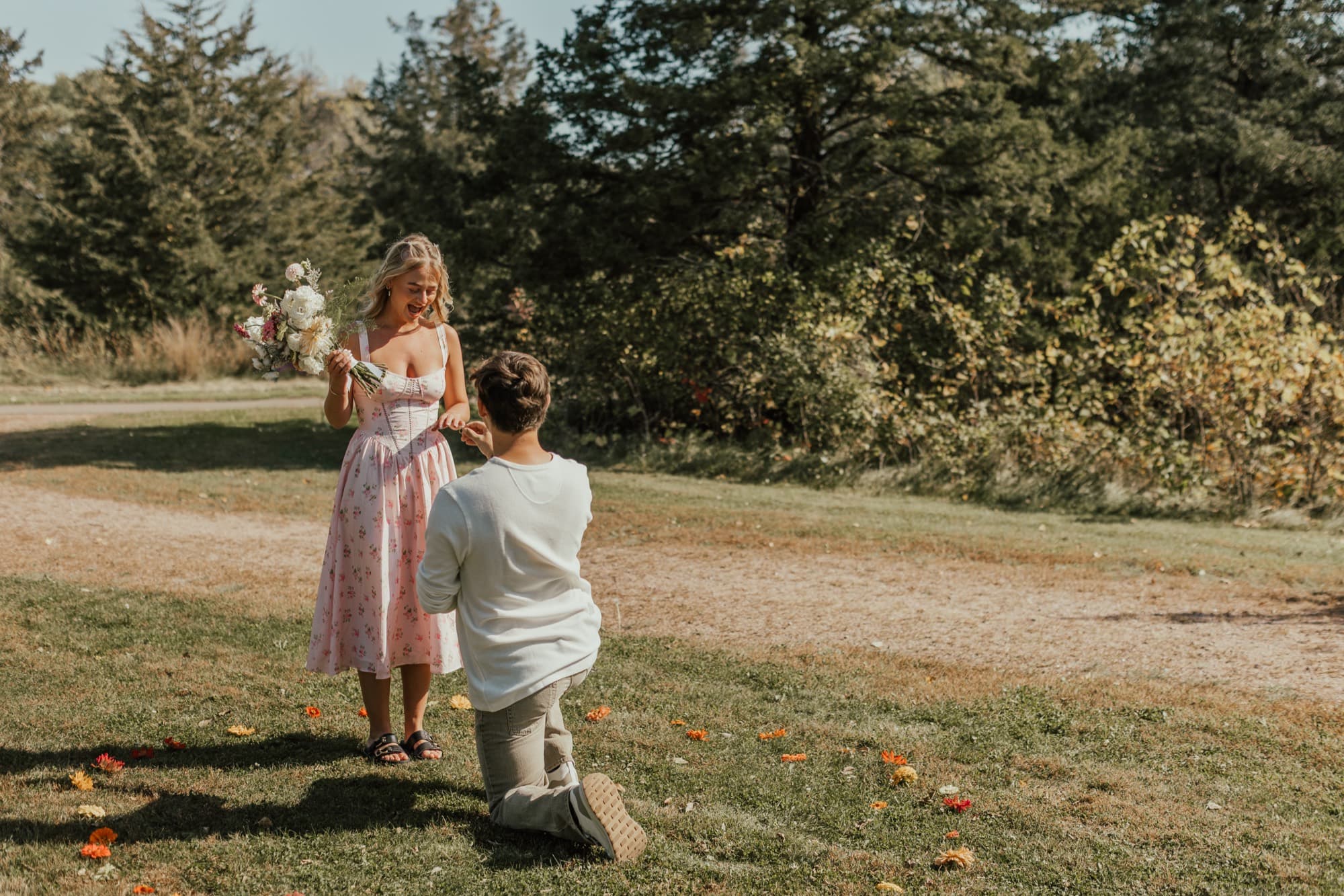 A man kneels to propose in an autumn field as the woman lifts her bouquet in surprise