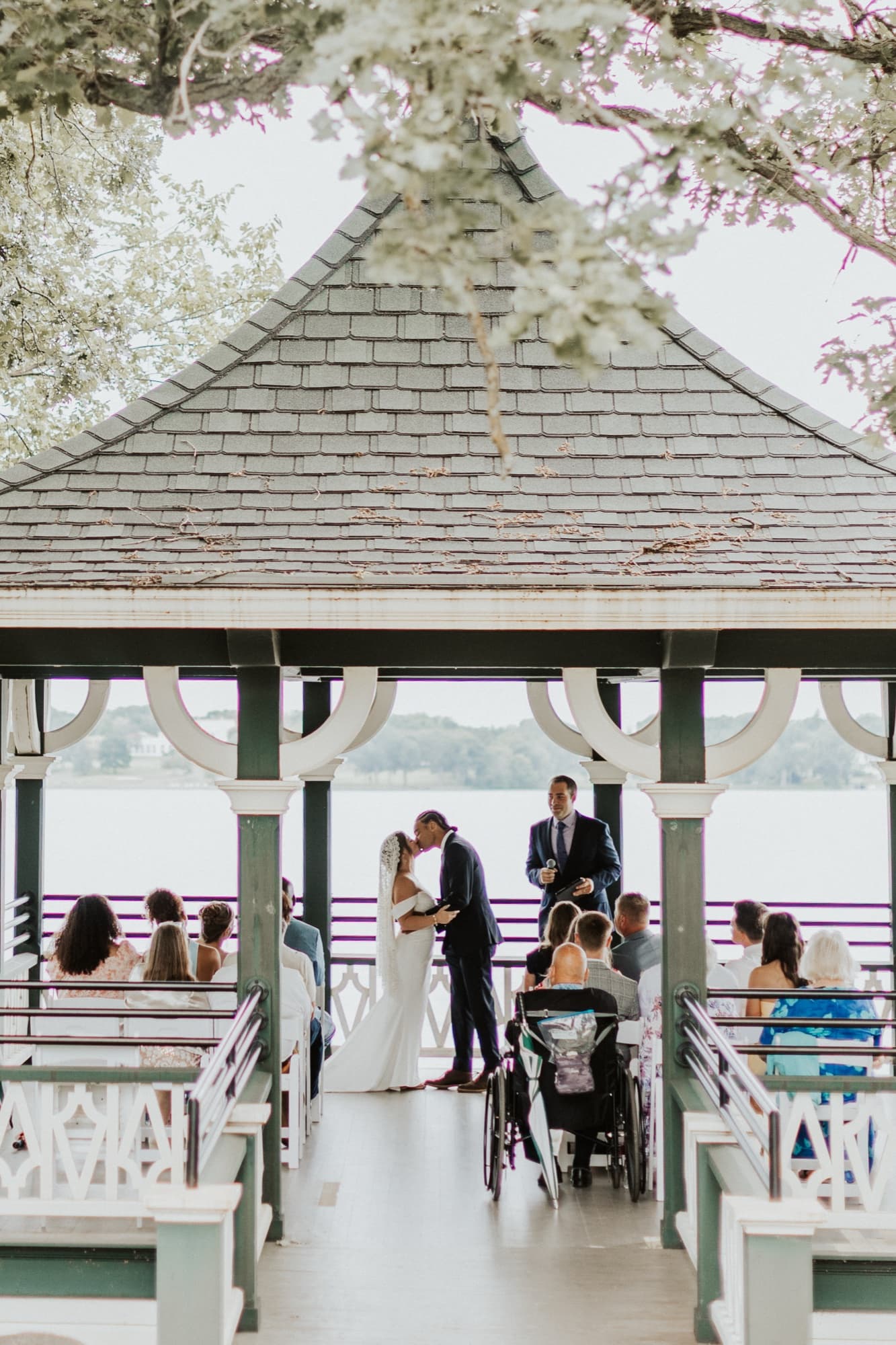 Nic and Tirzah kiss at the end of their ceremony in a white gazebo on the lake, guests seated on benches around them