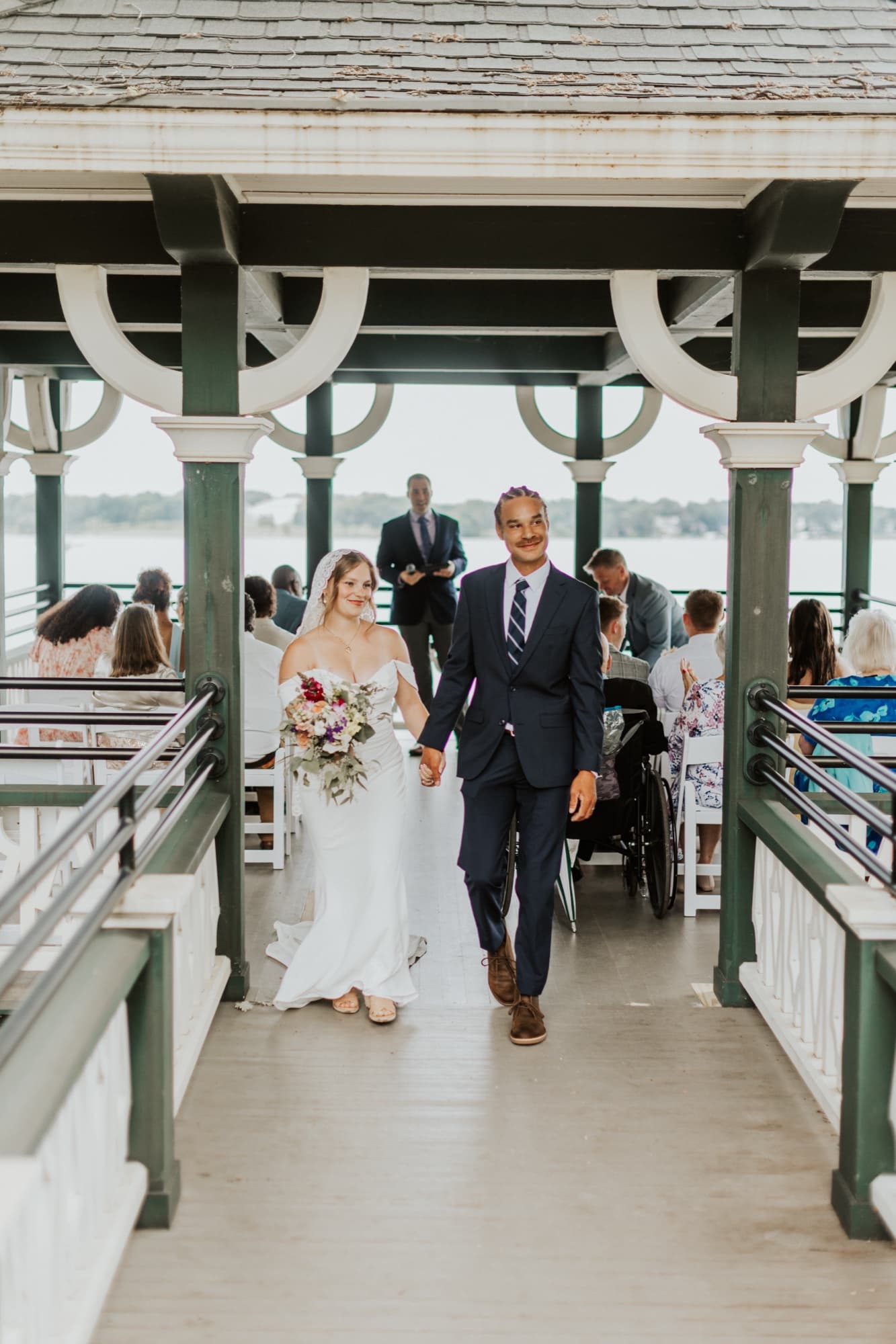 Nic and Tirzah walk back down the gazebo aisle hand in hand, smiling, as guests watch from the benches