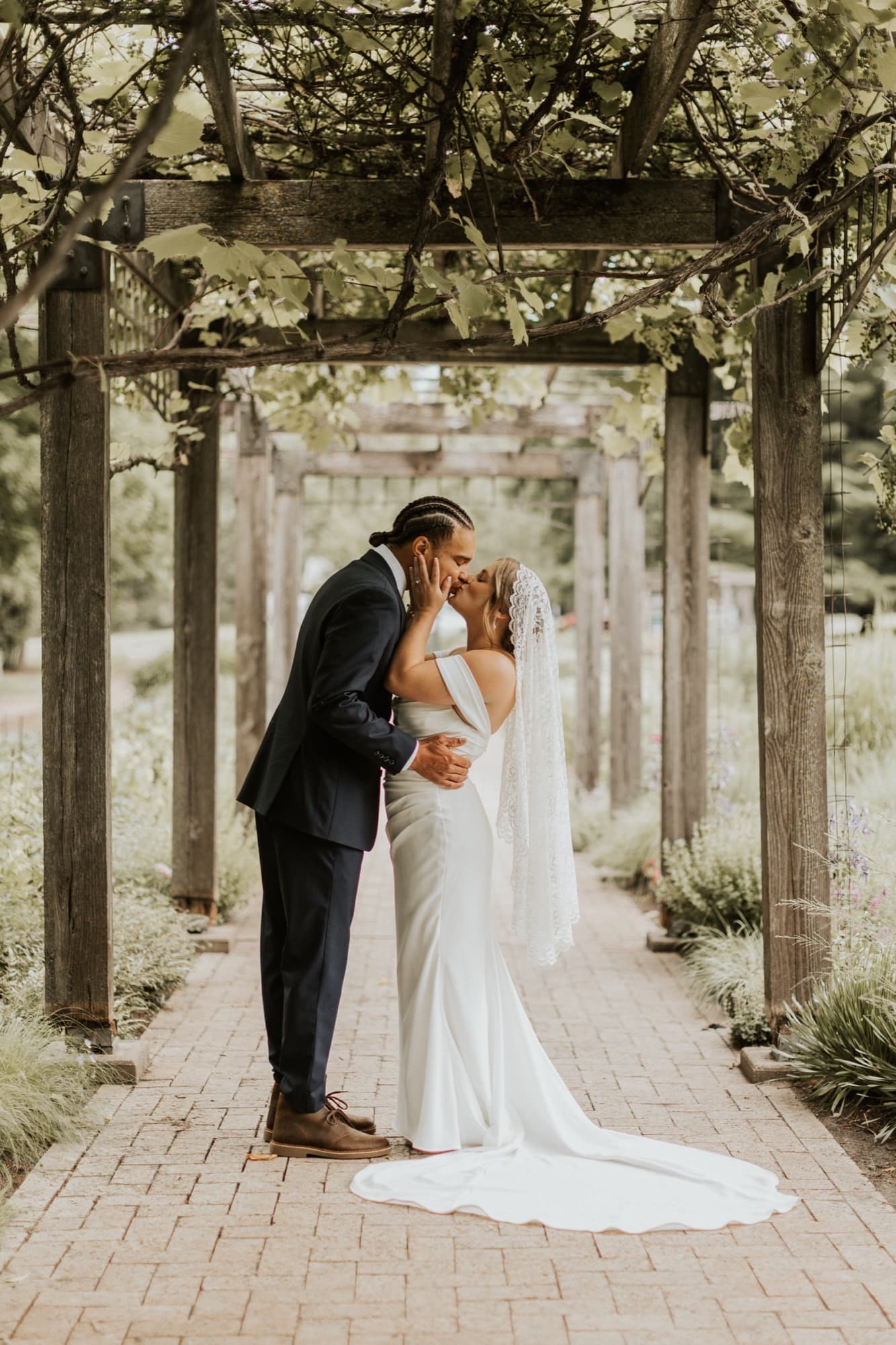 Nic and Tirzah kiss beneath a vine-covered wooden pergola on a brick path, her long train behind her