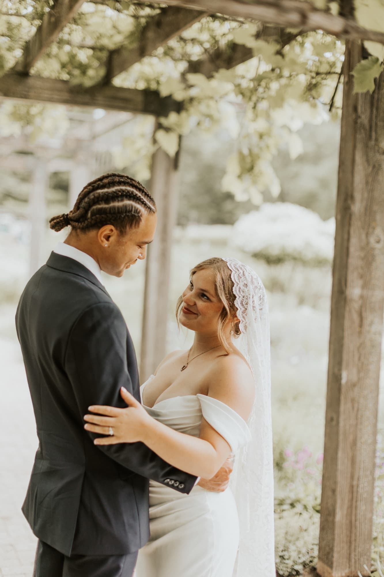 The bride, in a lace mantilla veil, looks up at the groom as he holds her beneath the pergola
